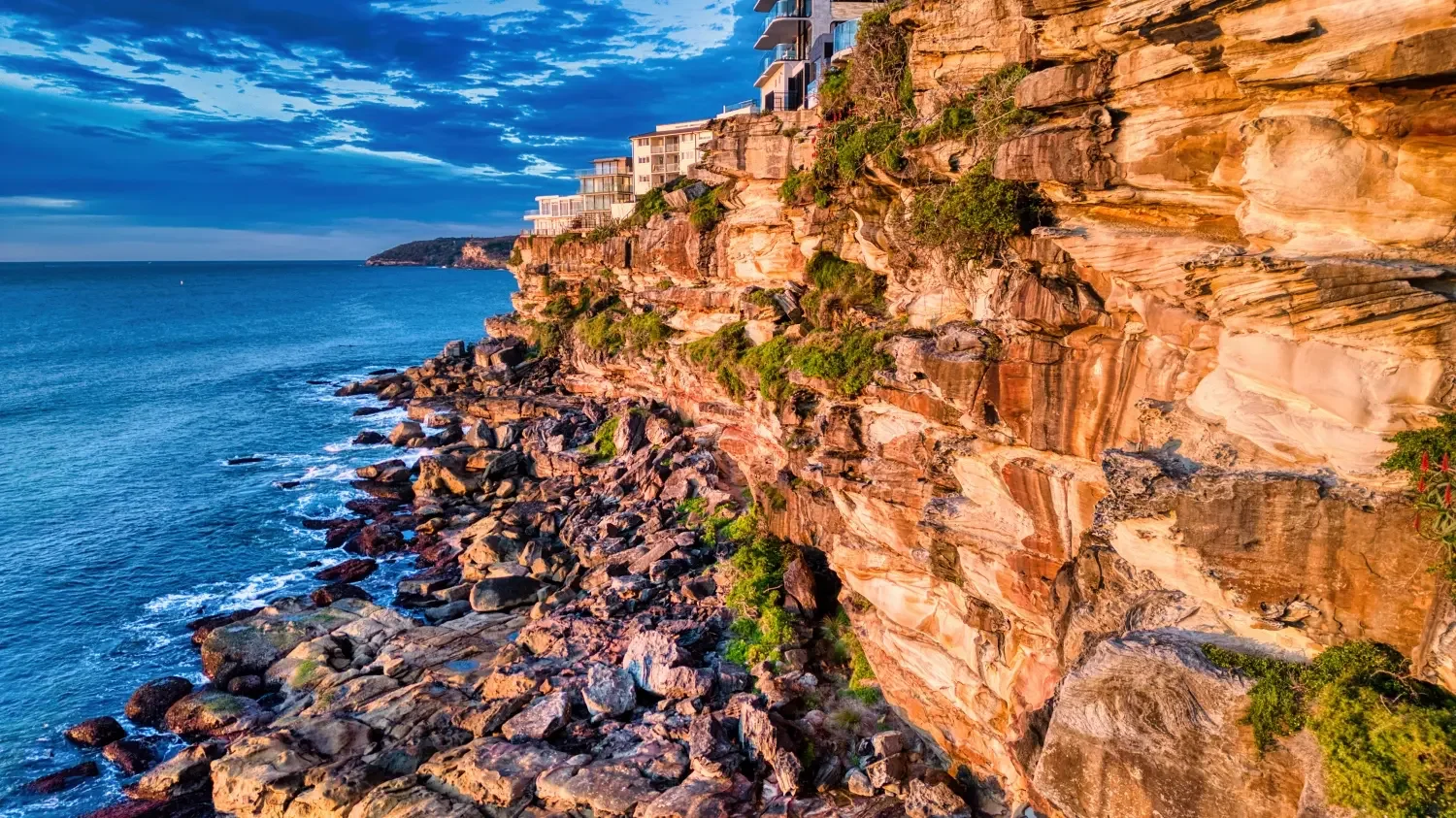 Cliffside buildings overlooking a rocky shoreline and ocean during sunset with partly cloudy sky.
