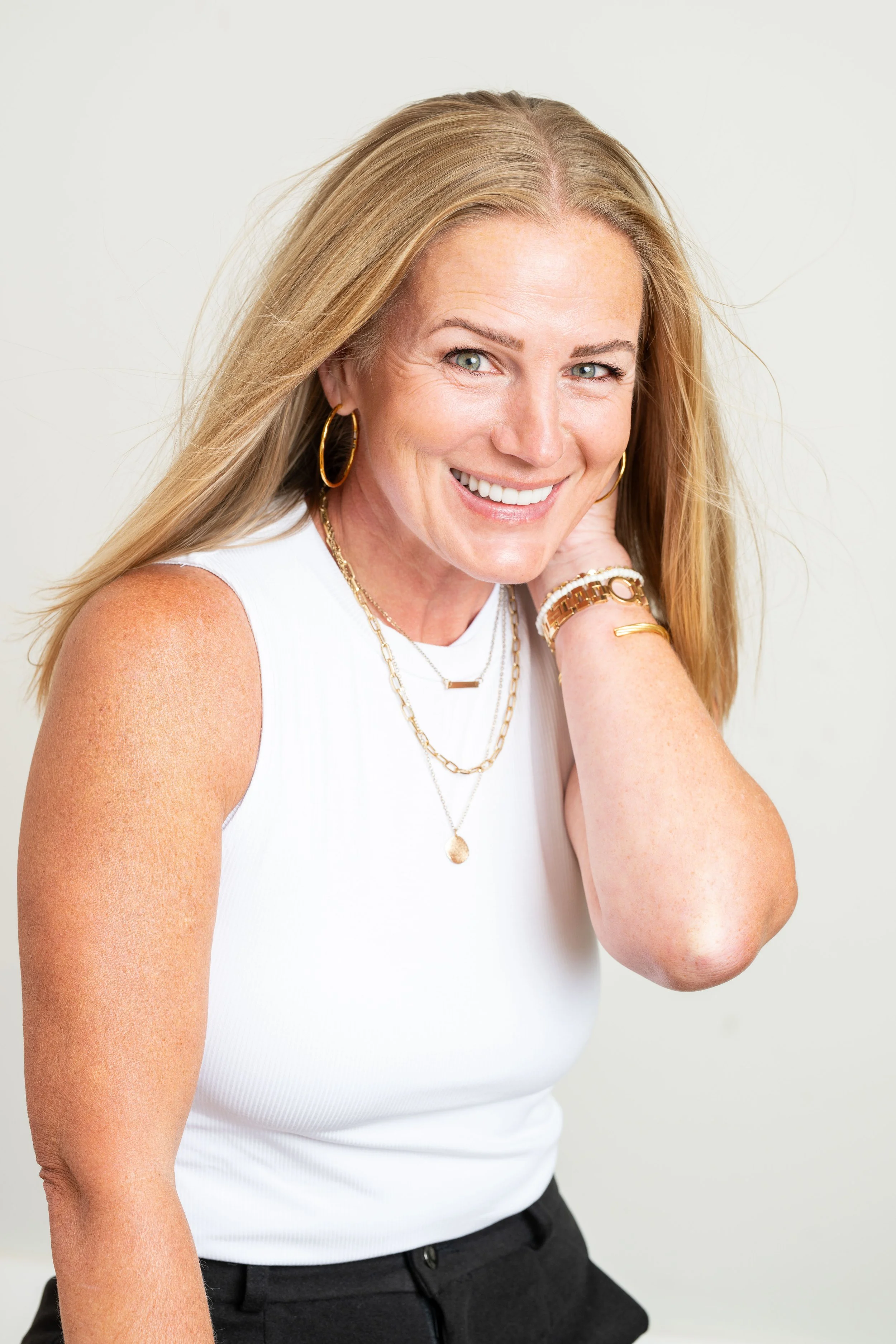 A woman with blonde hair wearing a white sleeveless top and gold jewelry, smiling and resting her hand on her neck against a plain white background.