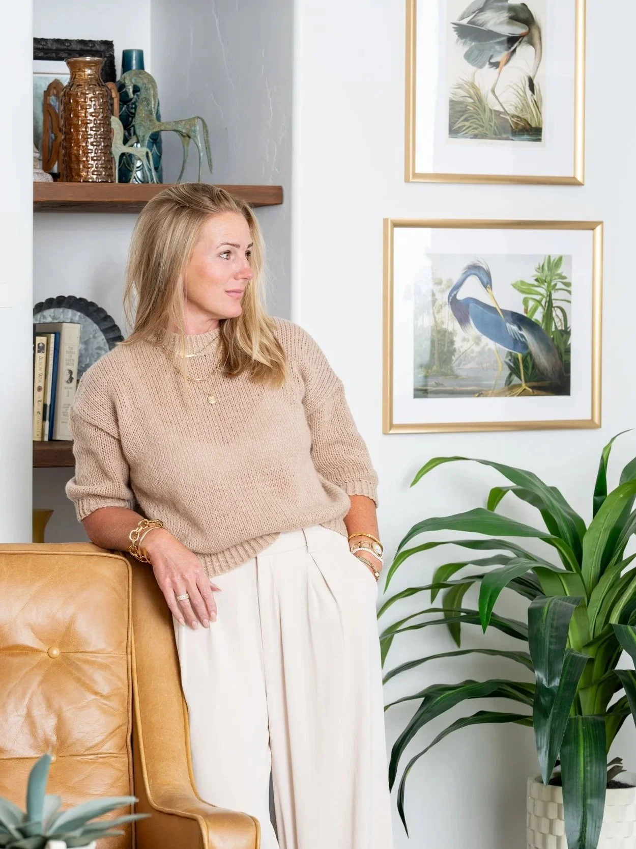 A woman with blonde hair wearing a beige sweater and white pants stands next to a tan leather chair in a decorated room, with framed bird artwork on the wall and a large green plant nearby.