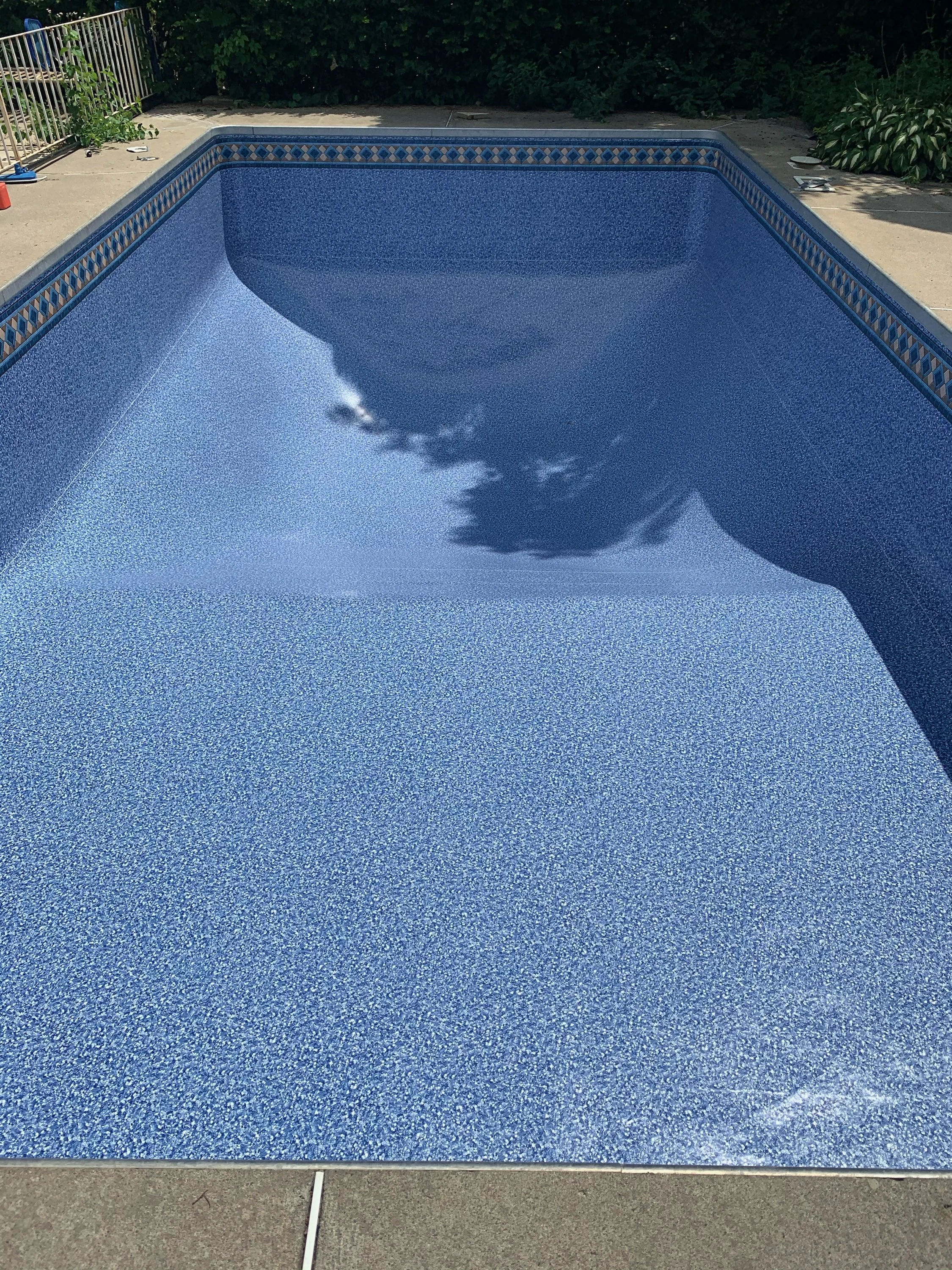 Empty swimming pool with blue mosaic tiles, reflection of trees on the water surface, surrounded by concrete patio and greenery.