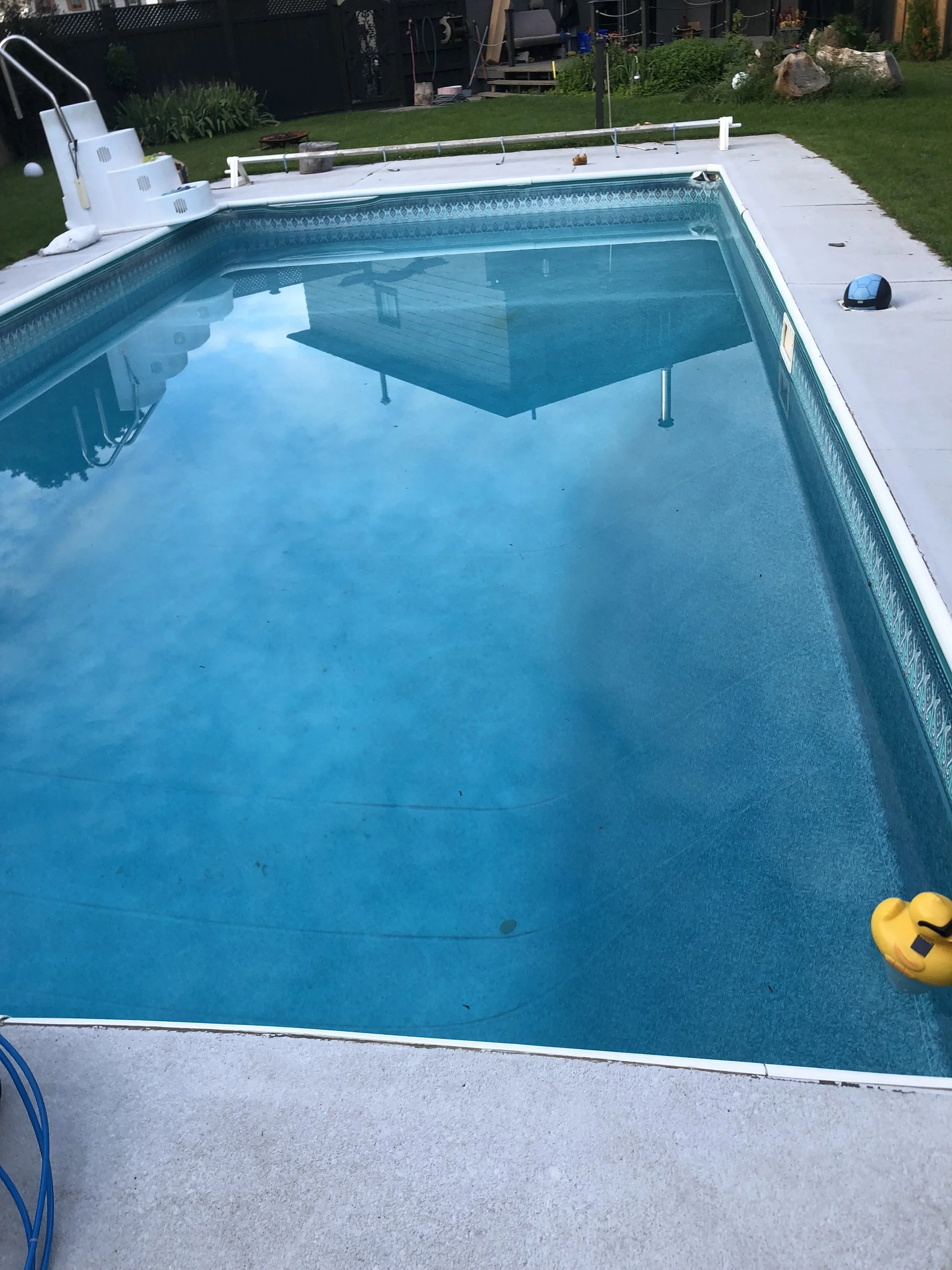 Empty in-ground swimming pool with blue water, partially covered pool liner, yellow rubber duck float, and surrounding concrete patio, back yard with green grass, plants, rocks, and garden furniture.