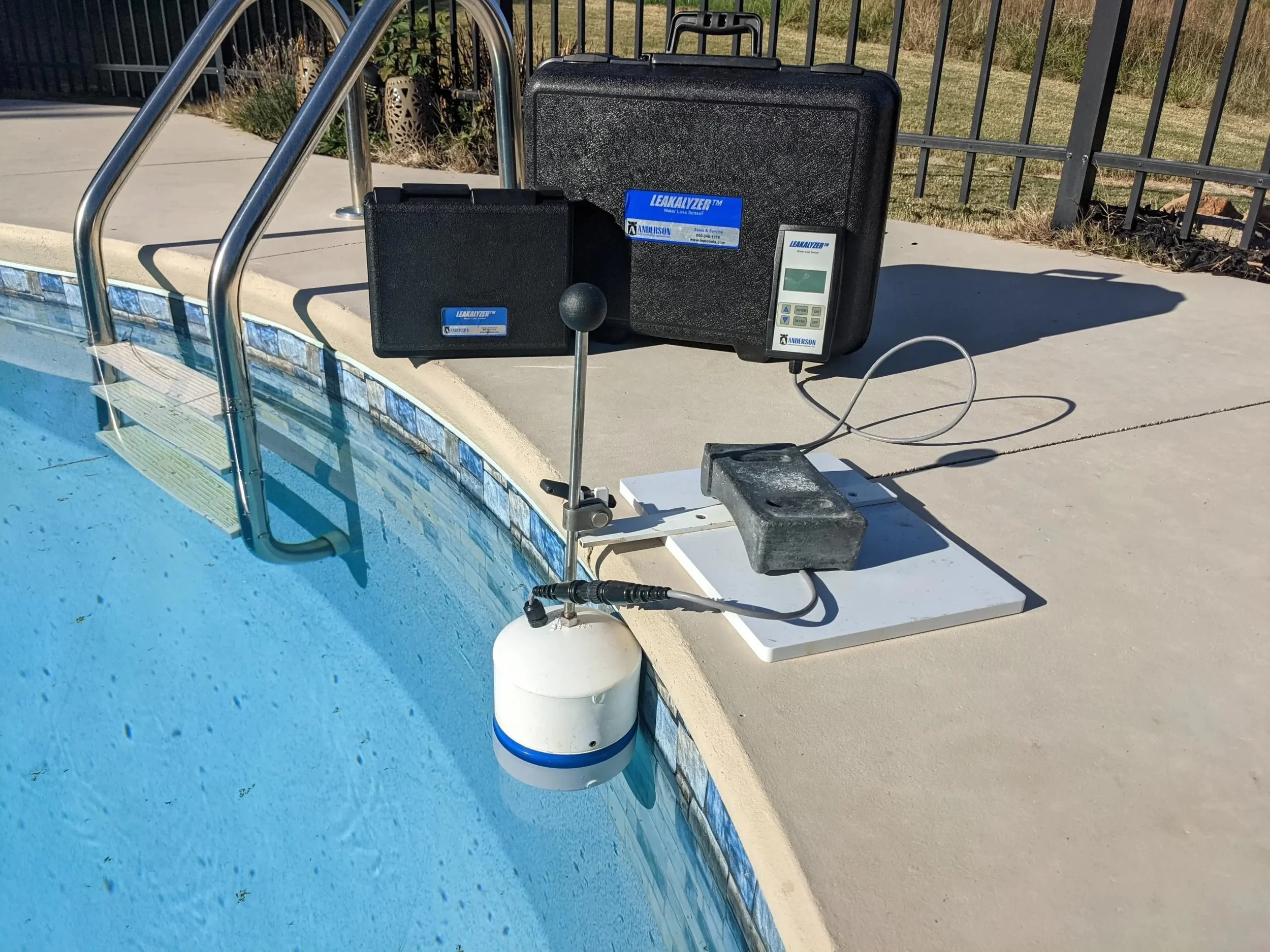 Pool testing equipment on the pool deck including a device with a white float, digital measuring units, and cases with labels near the poolside.