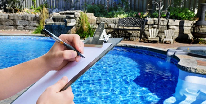 Person writing on a clipboard near a swimming pool with a stone wall and garden in the background.