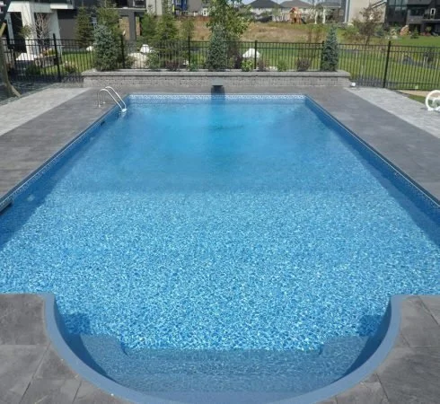 An outdoor swimming pool with blue water, surrounded by a grey stone patio and a black metal fence. There are residential buildings and green grassy areas in the background.