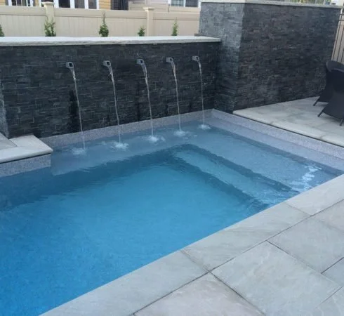 Infinity pool with four water features over a dark gray stone wall, adjacent to a concrete patio, in a backyard.