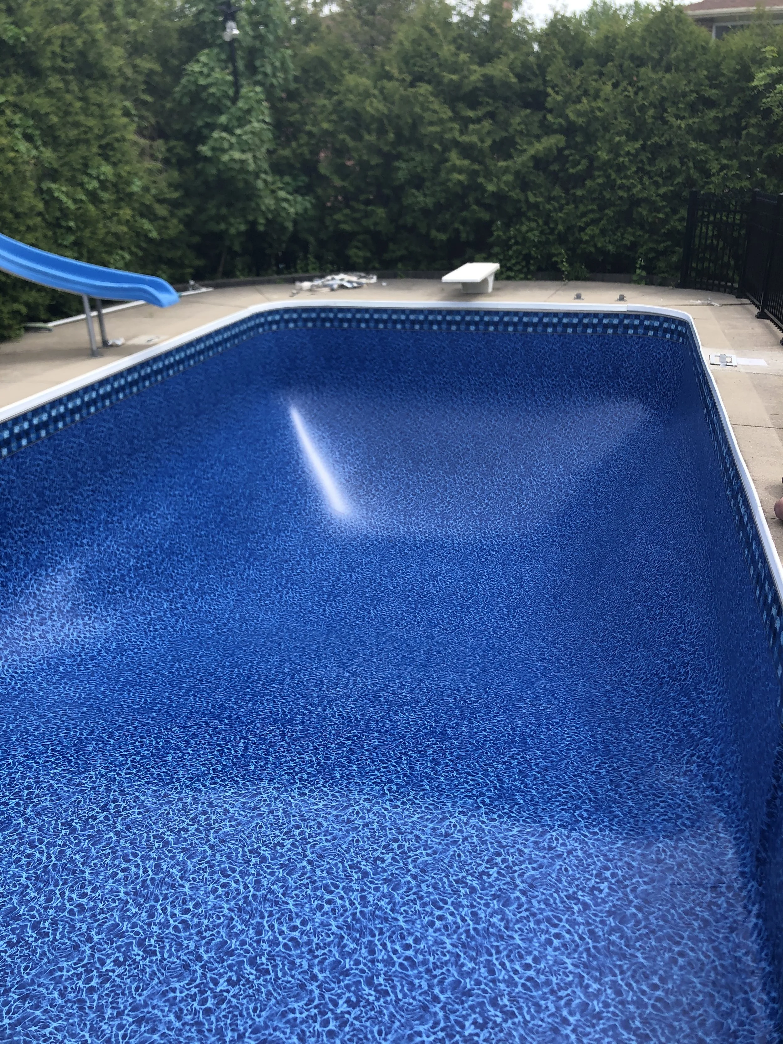 Empty blue swimming pool with water slide and green trees in the background.