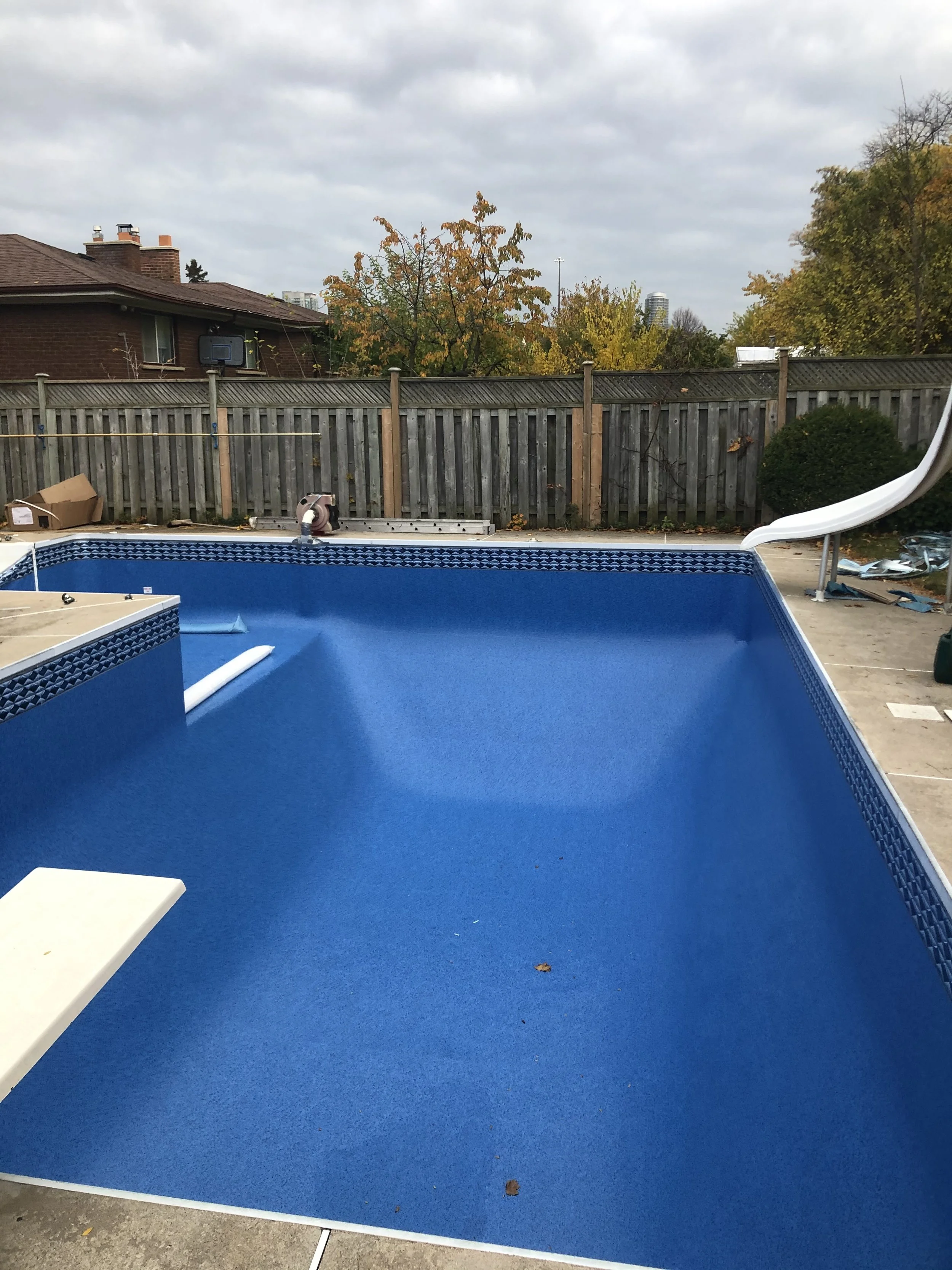 An empty swimming pool under construction with a blue interior and a slide on the right side, surrounded by a concrete deck and a wooden fence, with trees and houses in the background.