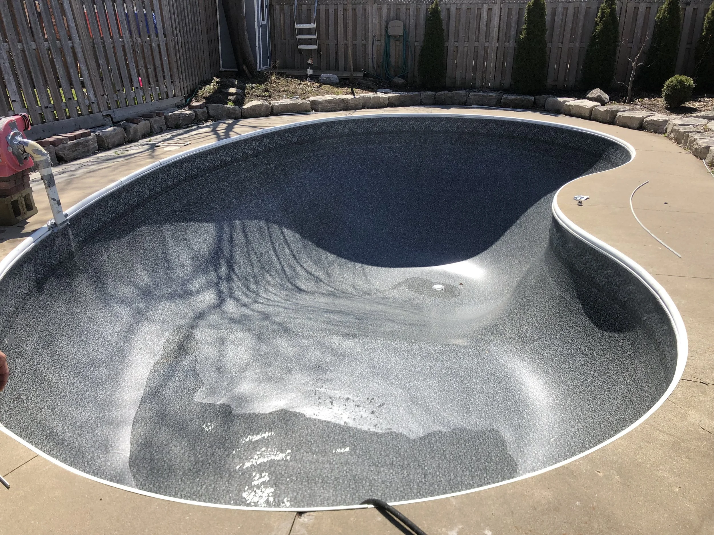 Empty kidney-shaped swimming pool with black interior and white border, situated outdoors on a concrete patio, surrounded by a wooden fence with plants and garden tools in the background.