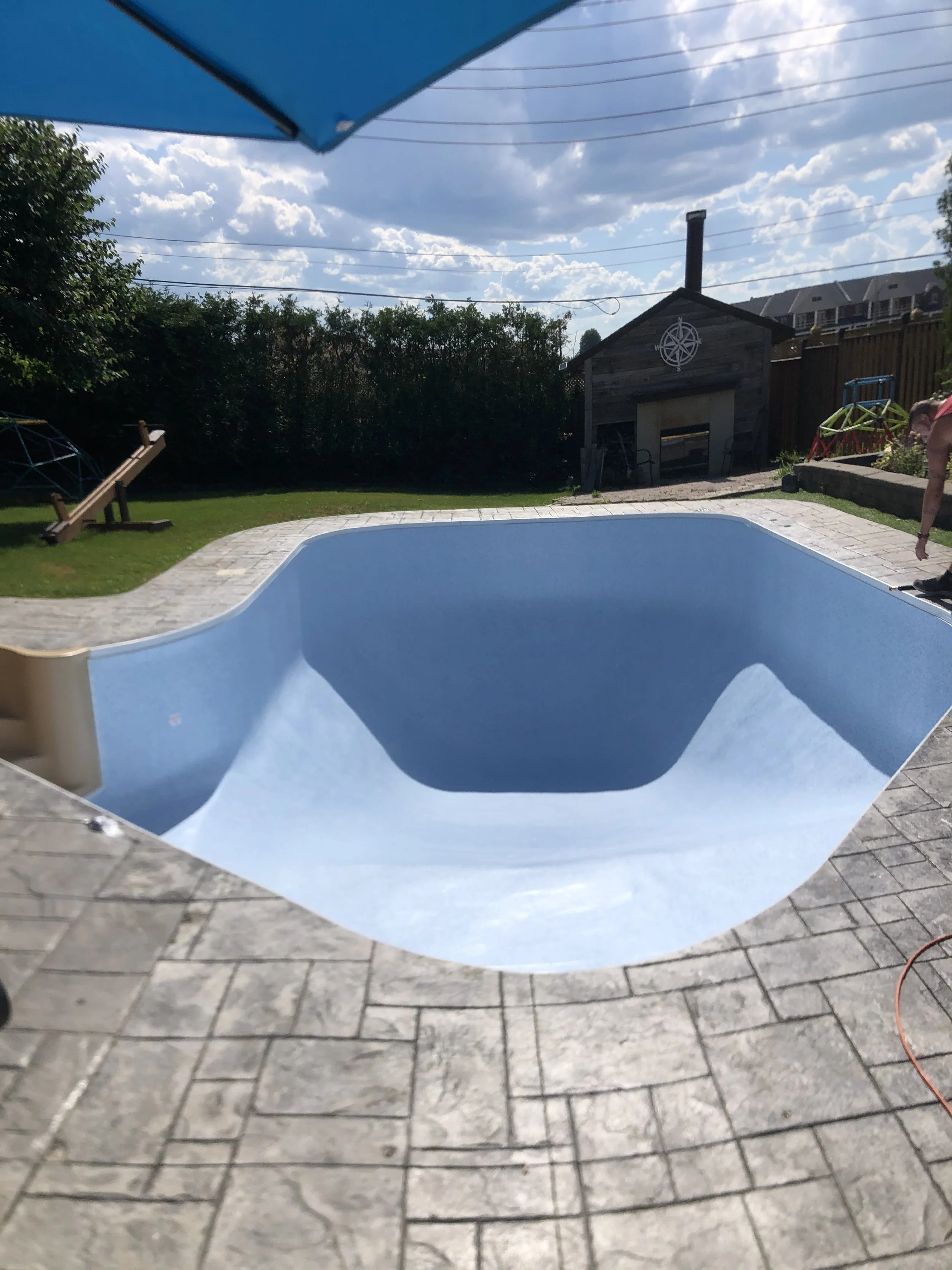Empty renovated backyard pool with a blue interior, surrounded by a stone patio, with a cloudy sky overhead and a small wooden shed, a slide on the grass, and some play equipment in the background.