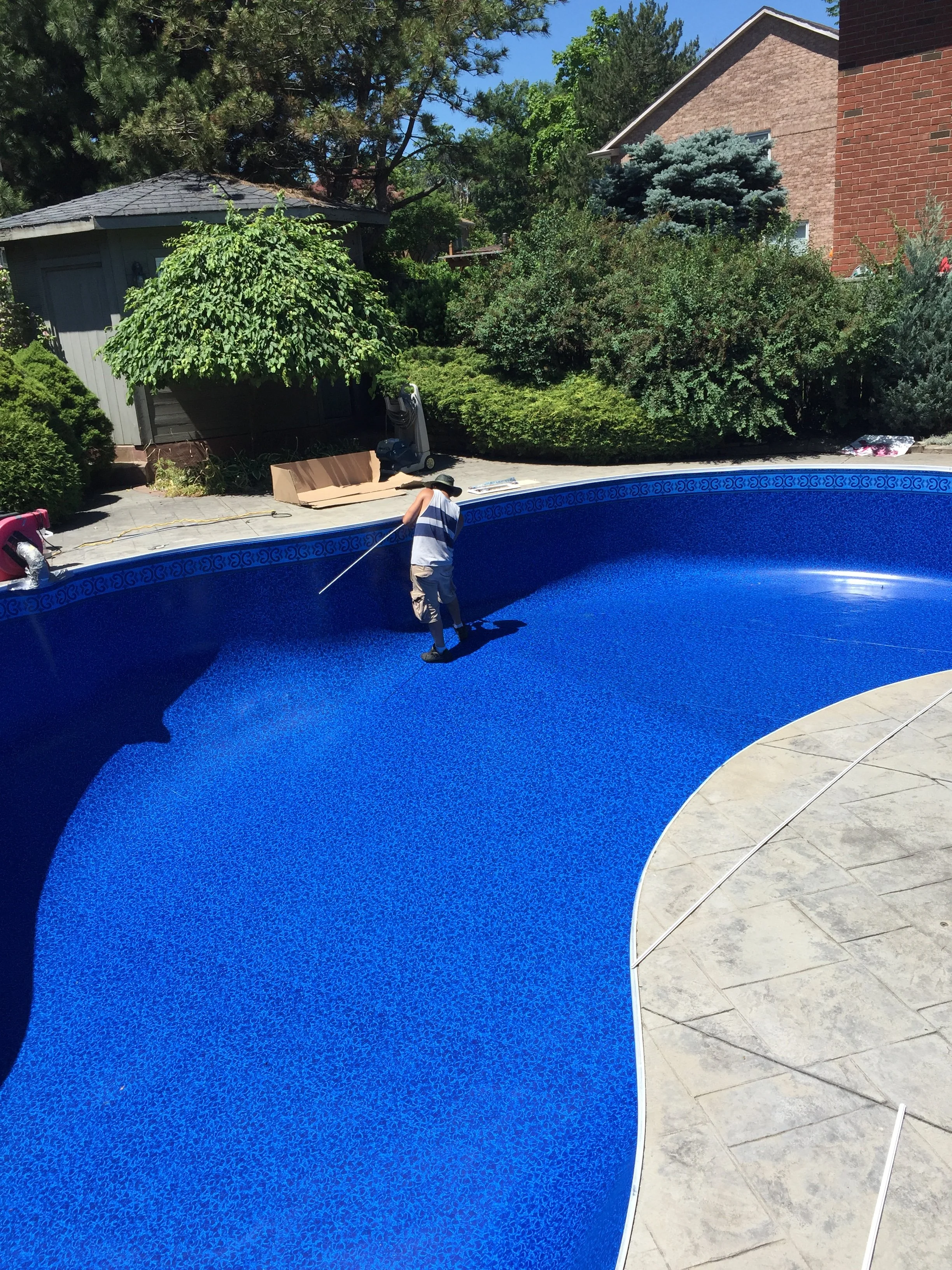 A person cleaning the inside of an empty swimming pool with a long-handled brush or tool in a backyard on a sunny day.