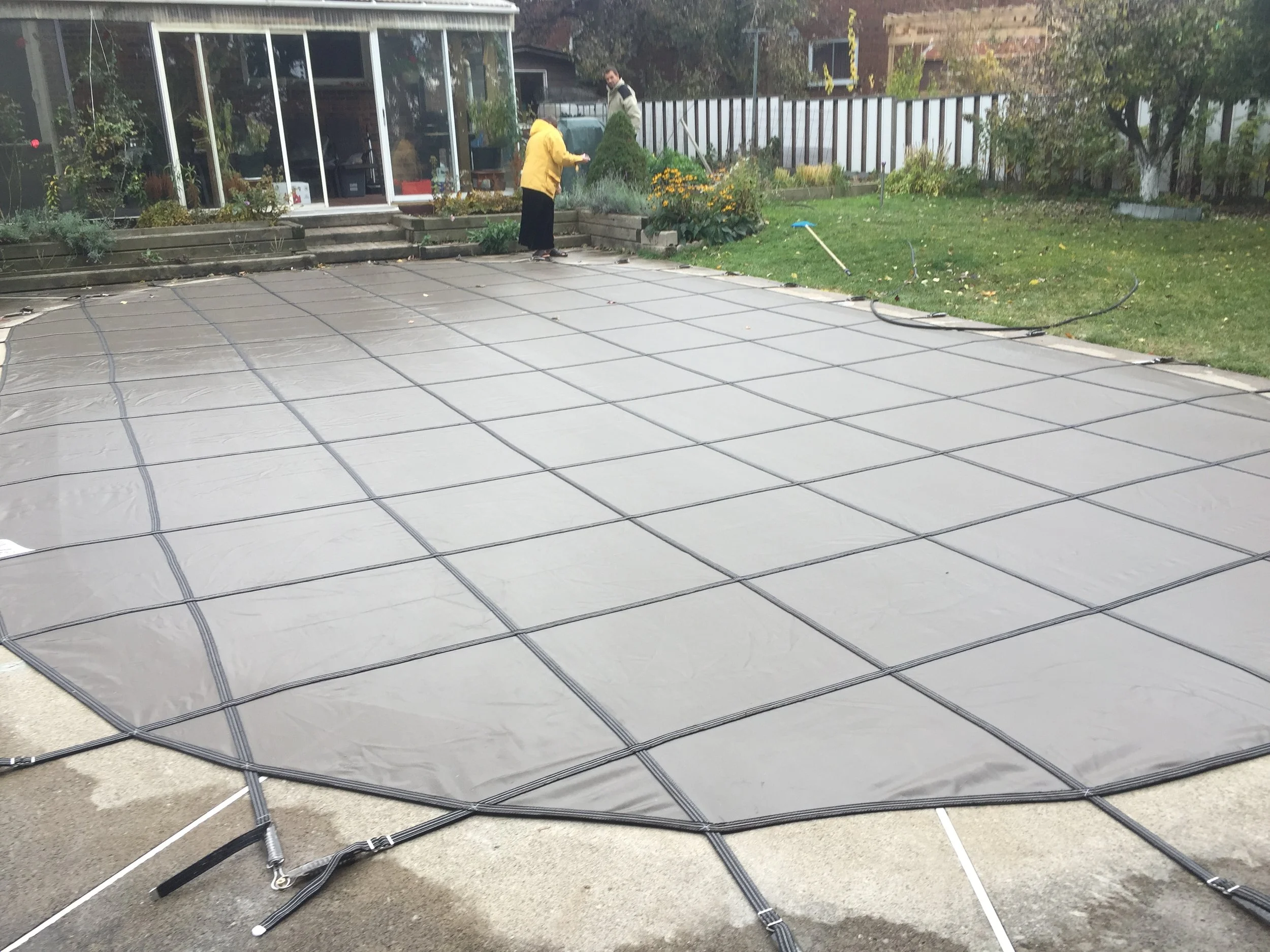 People installing a concrete or stone surface using a grid pattern in a backyard, with a garden and a glass external room in the background.