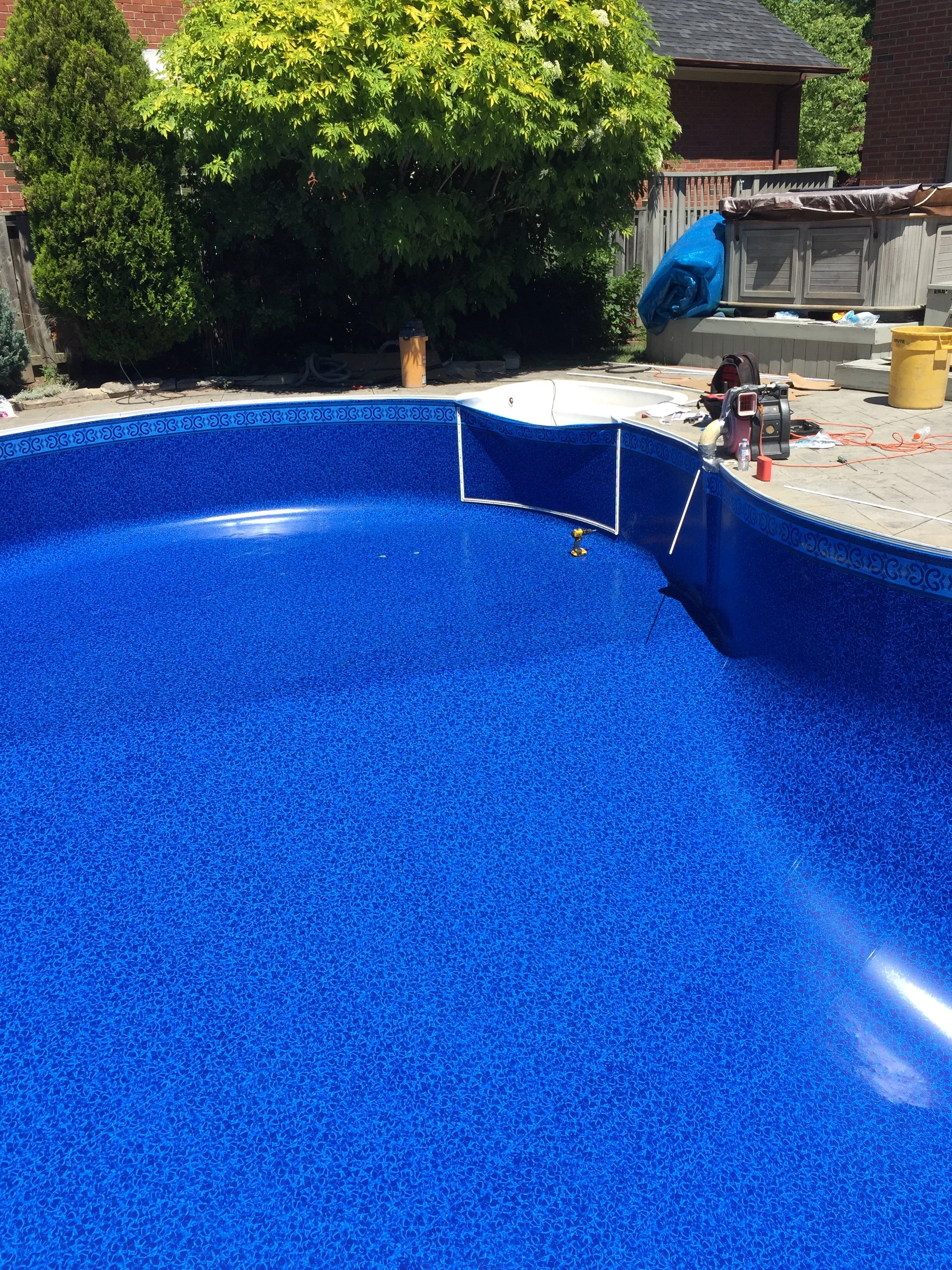 An empty in-ground swimming pool with a bright blue interior, surrounded by a patio area with pool equipment and tools, with a green tree and fence in the background.