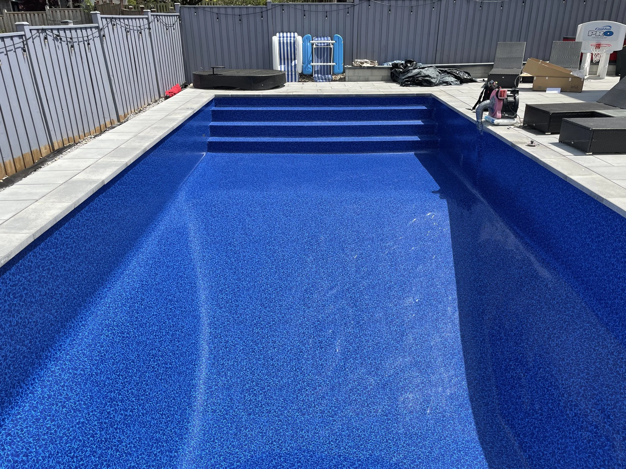 Blue swimming pool with built-in steps, surrounded by a tiled deck, with pool cleaning equipment and outdoor furniture nearby.