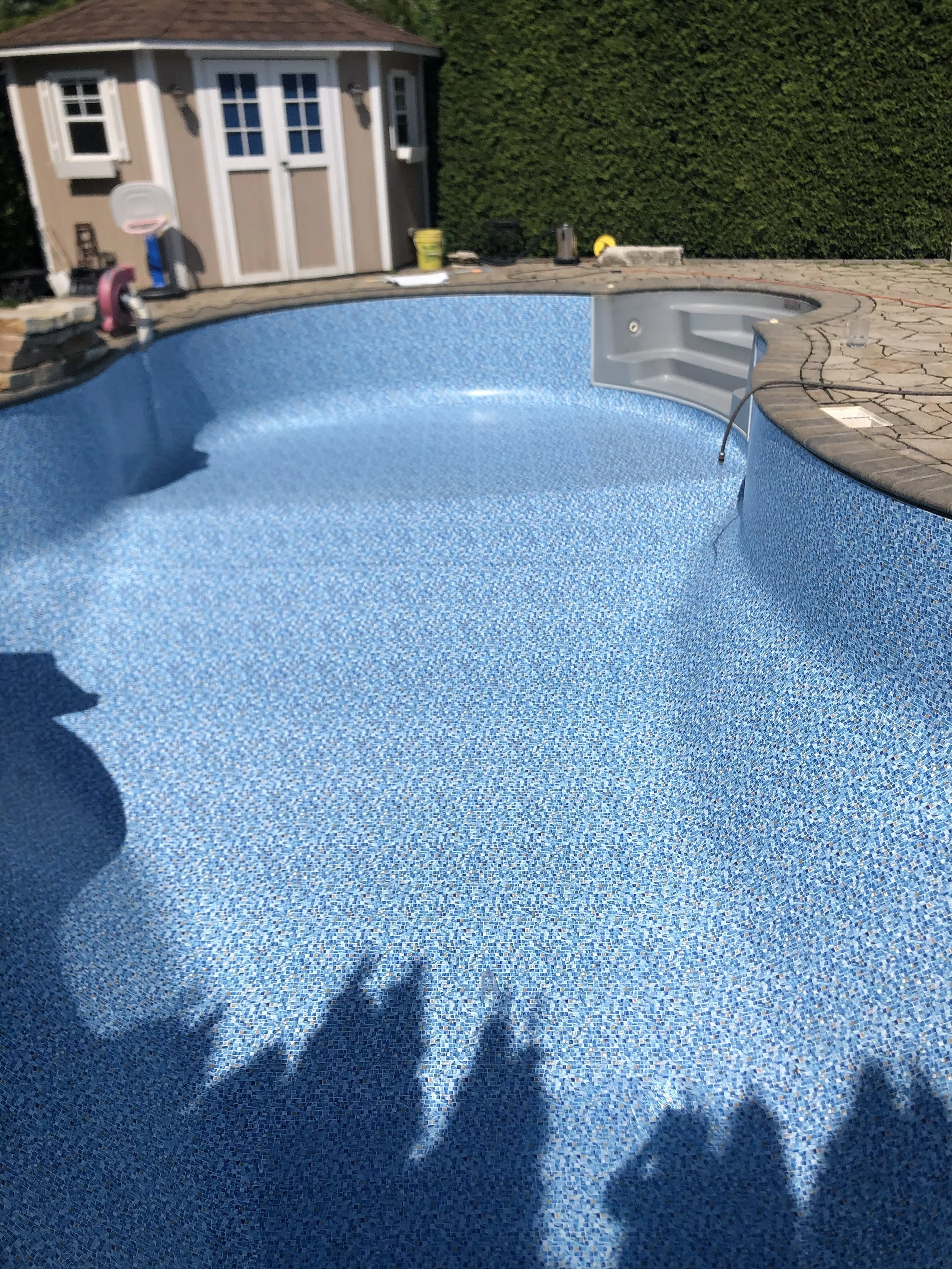 Empty in-ground swimming pool with blue mosaic tiles, surrounded by a stone deck, and a small shed in the background.