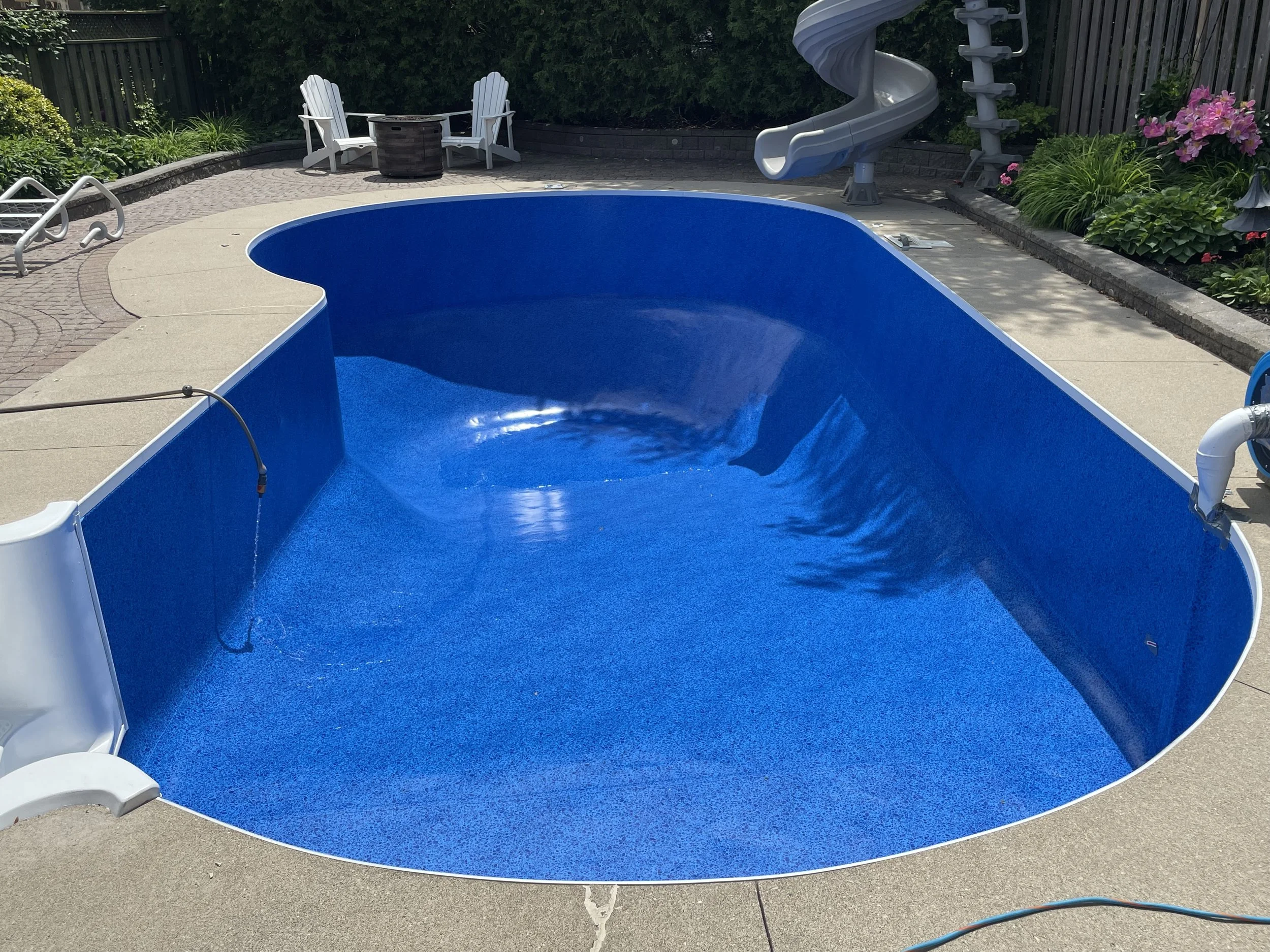 Empty blue in-ground swimming pool with a water slide, pool cover roller, and lounge chairs on a concrete deck, surrounded by a garden with flowers and plants.