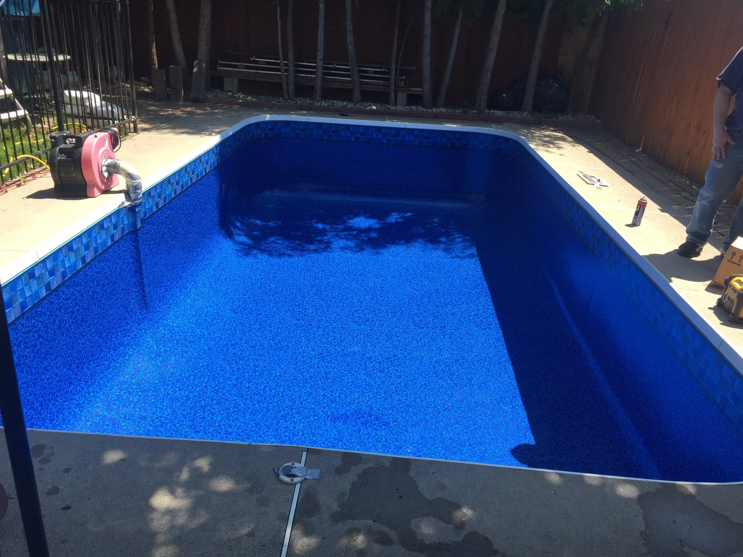 Blue in-ground swimming pool under construction with a partially tiled edge, surrounded by a concrete deck, fence, and tools.