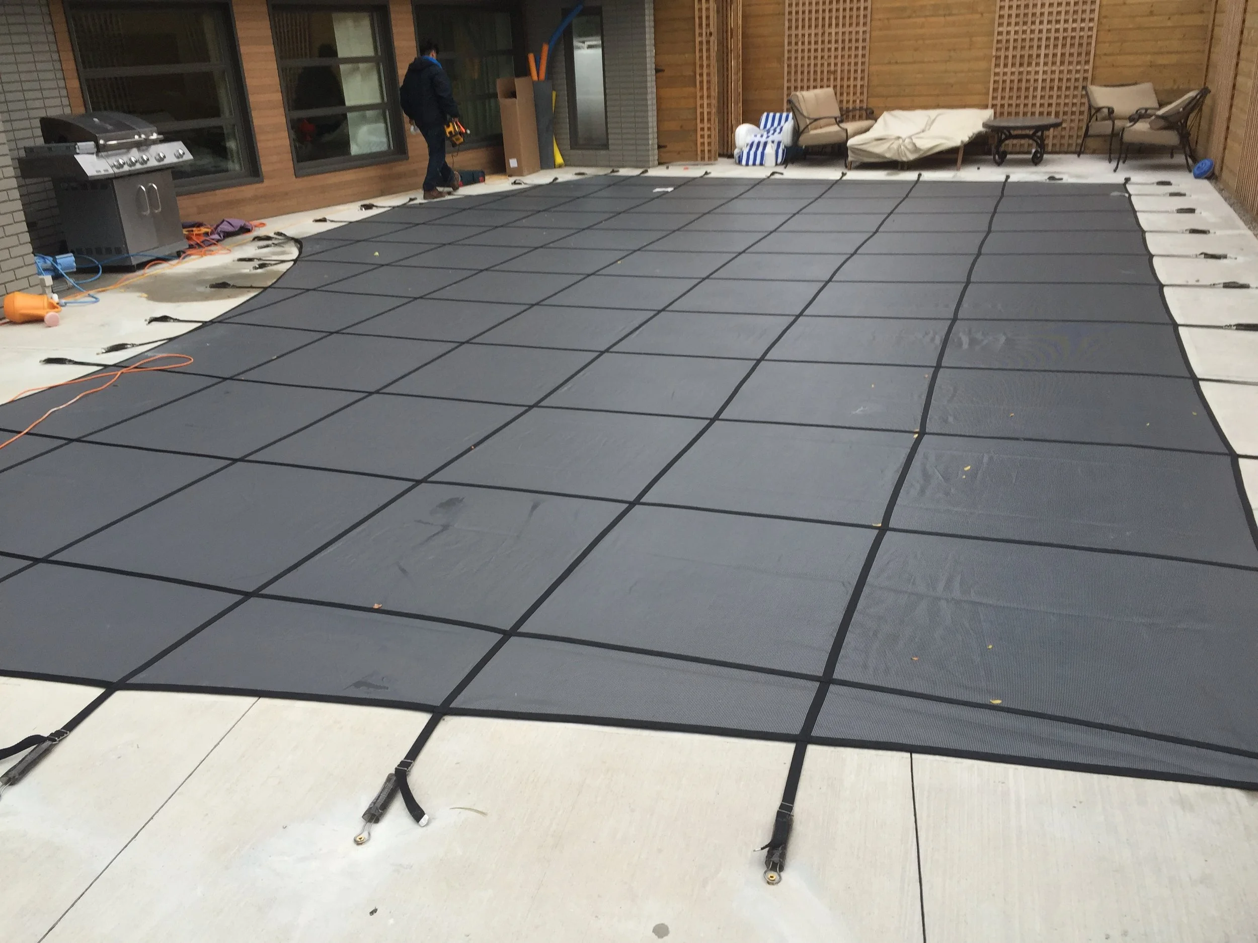 A backyard patio area with a black safety net laid out over a concrete surface, likely for a pool. There are lounge chairs, a small table, and some cushions against a wooden fence. A person is working near a sliding glass door on the left, with tools