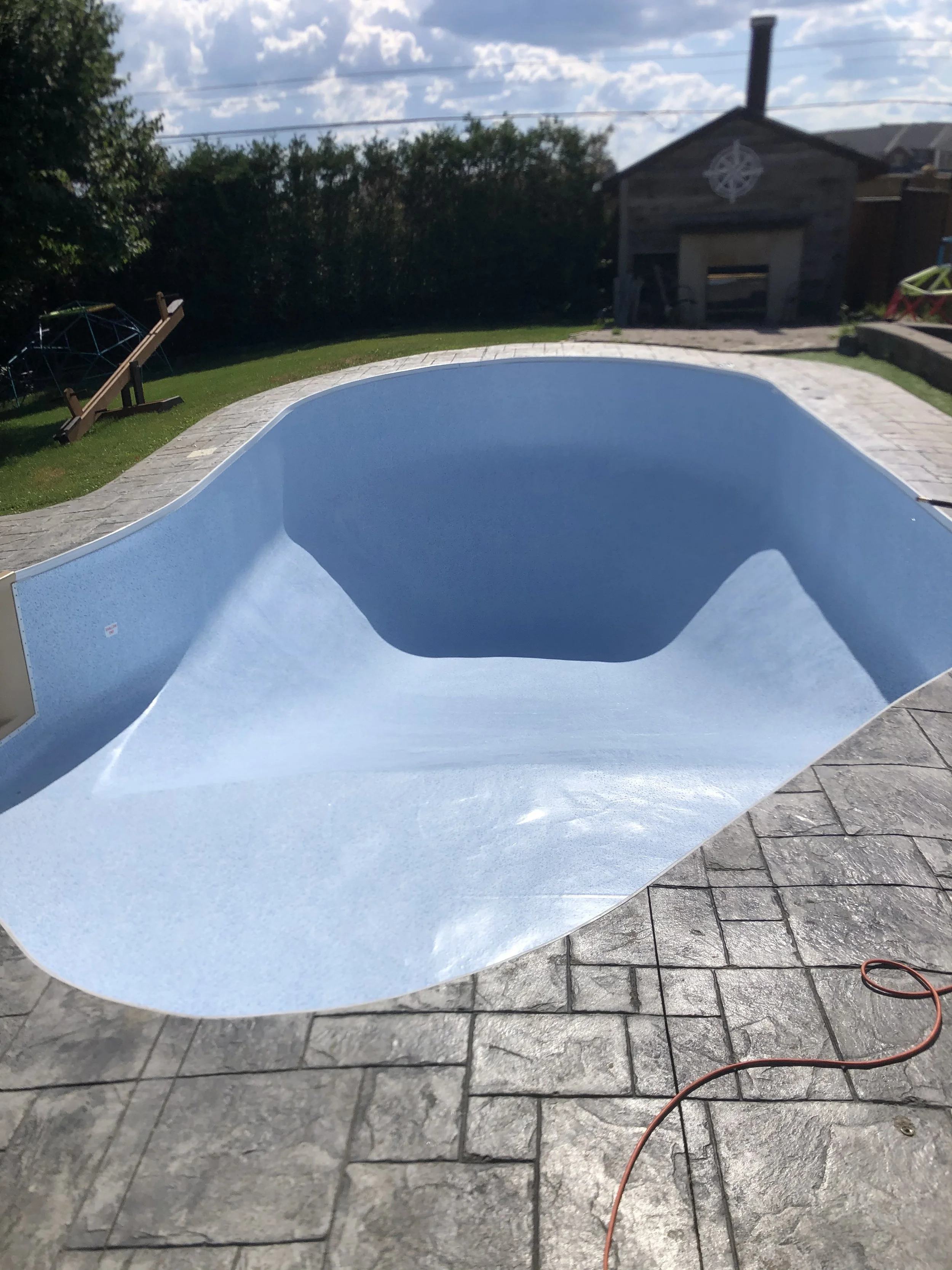 Empty swimming pool with a blue interior on a stone patio, backyard with a shed, lawn, trees, and outdoor furniture in the background.