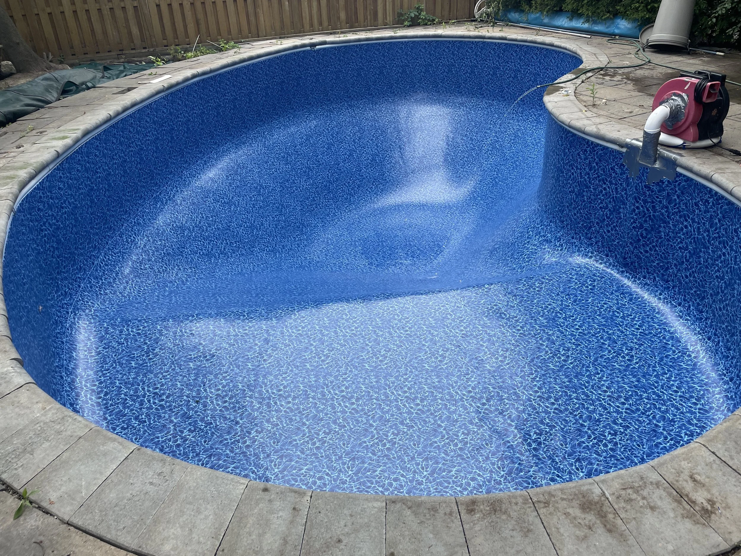 Empty backyard in-ground swimming pool with blue mosaic lining, surrounded by concrete paving, with a pool pump and filter system on the right side, and a wooden fence in the background.