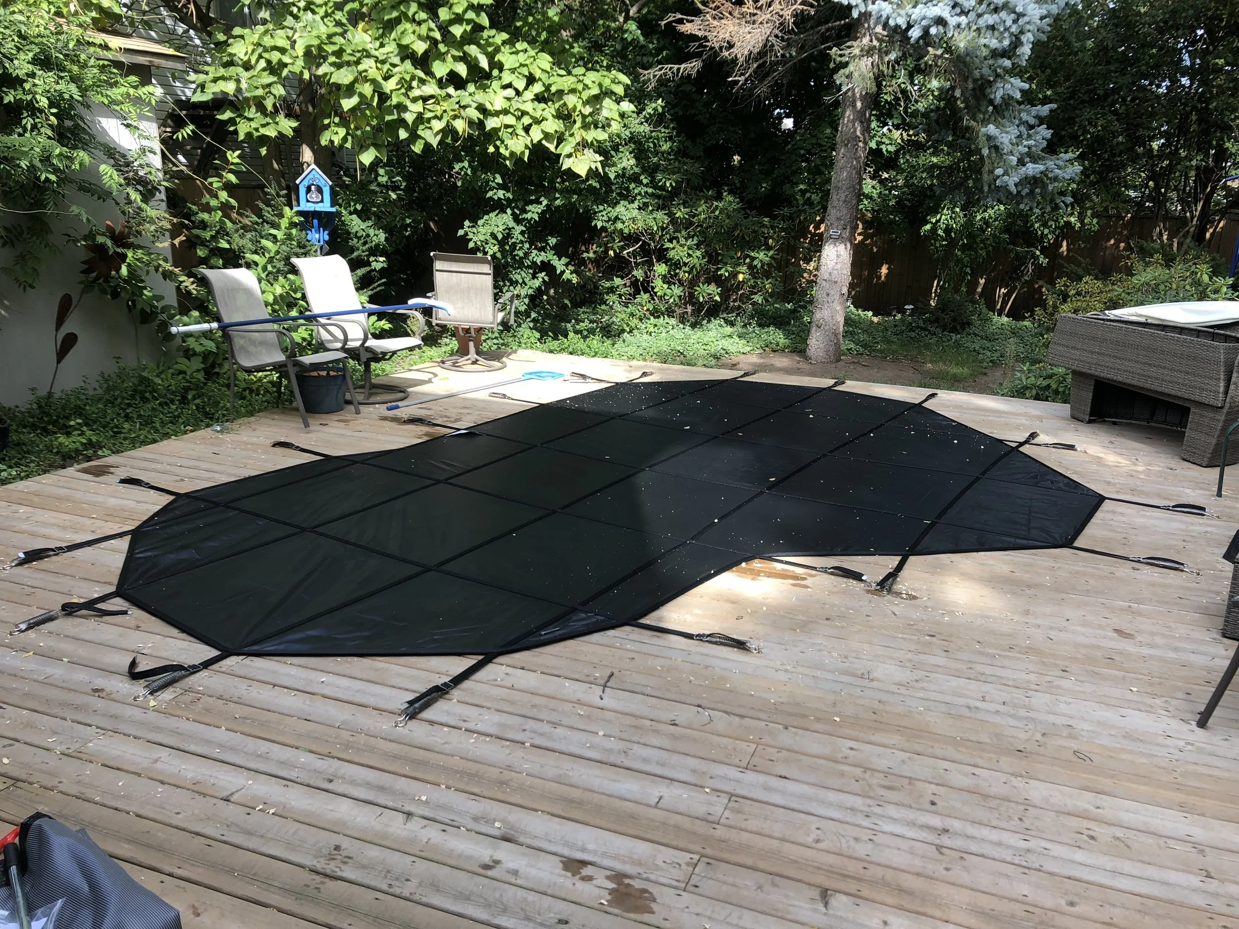 Sunshade canopy laid on a wooden deck in a backyard, surrounded by outdoor furniture and greenery.