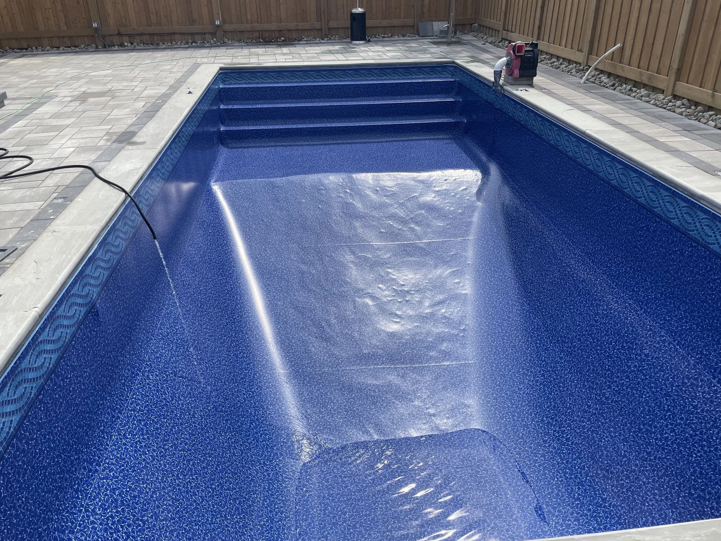 Empty in-ground swimming pool with blue tile interior, surrounded by a stone and concrete patio, fencing, and pool equipment.