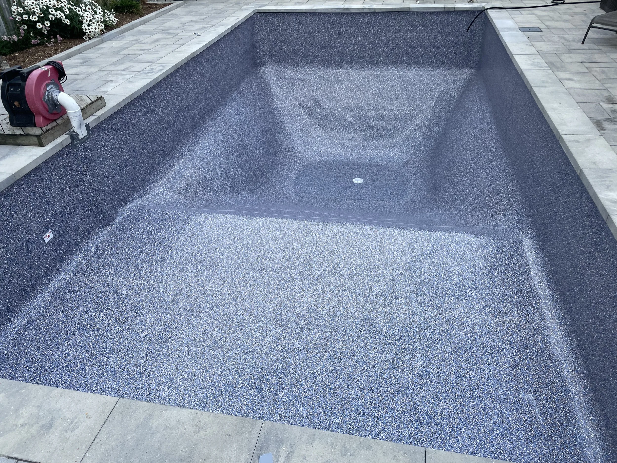 Empty in-ground swimming pool with a blue speckled interior, surrounded by a gray tiled deck, and some landscaping with white flowers in the background.