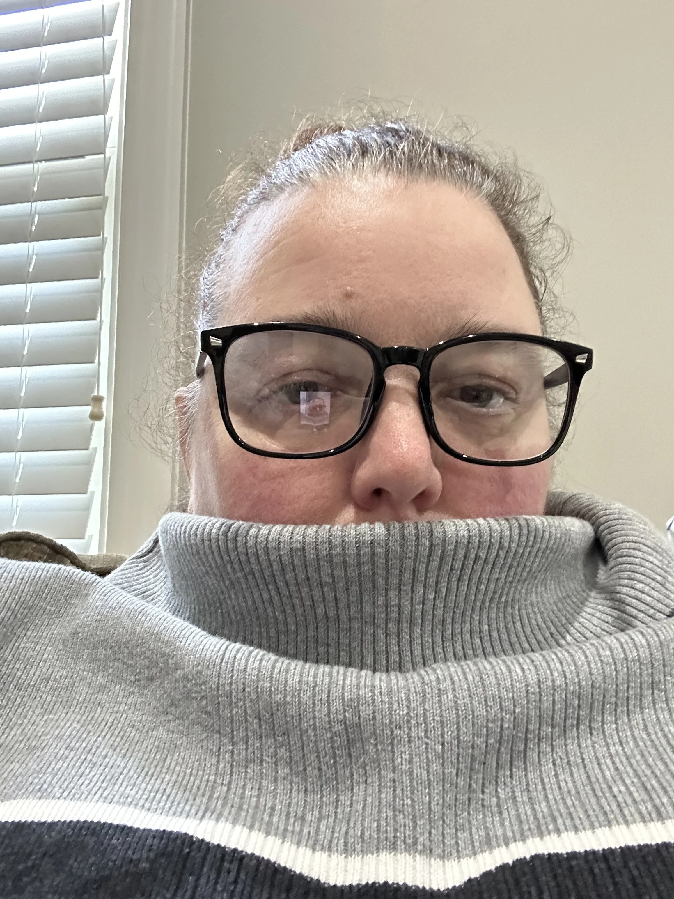 A person wearing black glasses and a gray sweater with the collar pulled up over their mouth, sitting indoors near a window with white blinds.