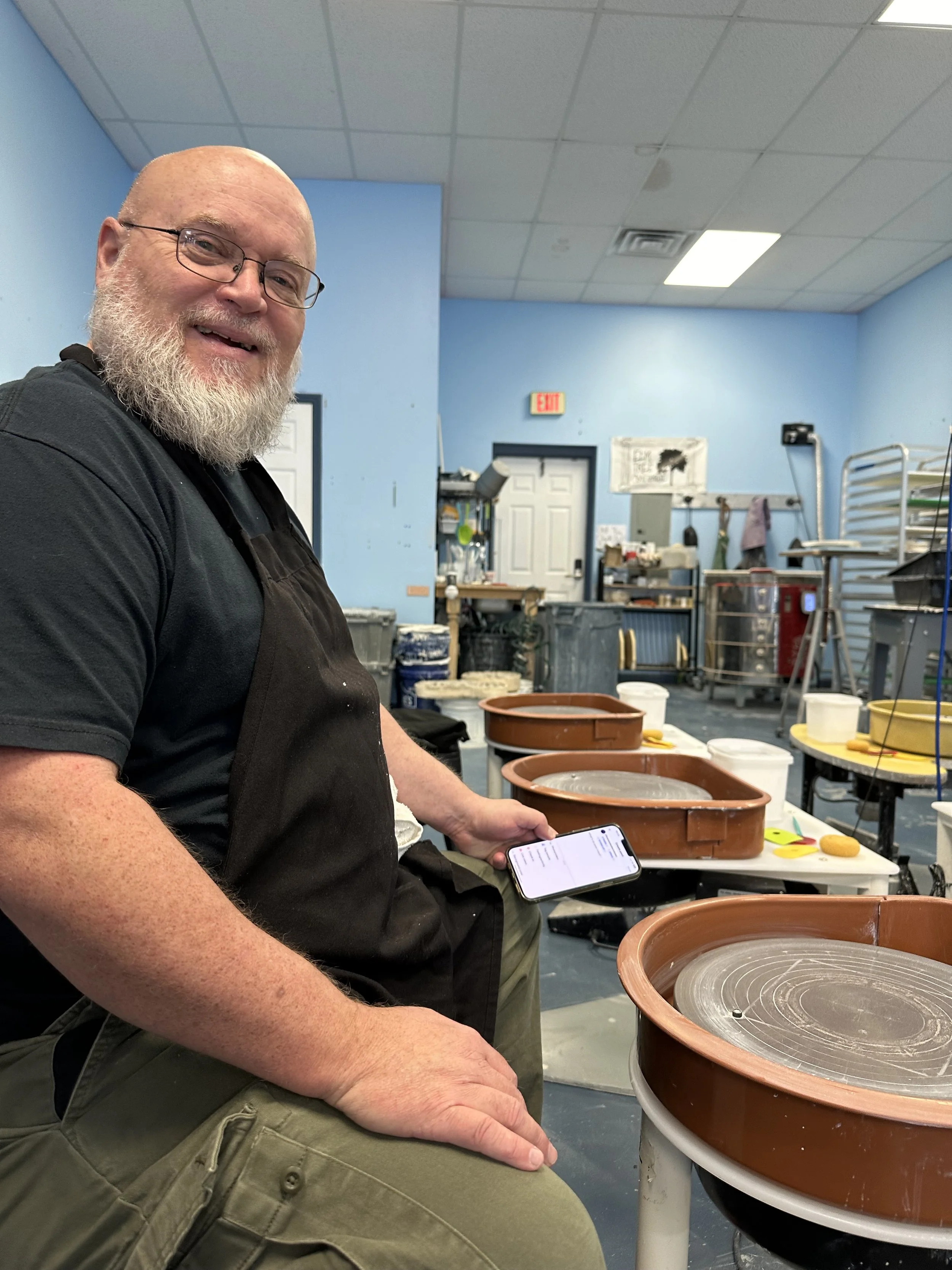 A smiling man with a beard and glasses, sitting in a ceramics studio surrounded by pottery molds and equipment, holding a smartphone.