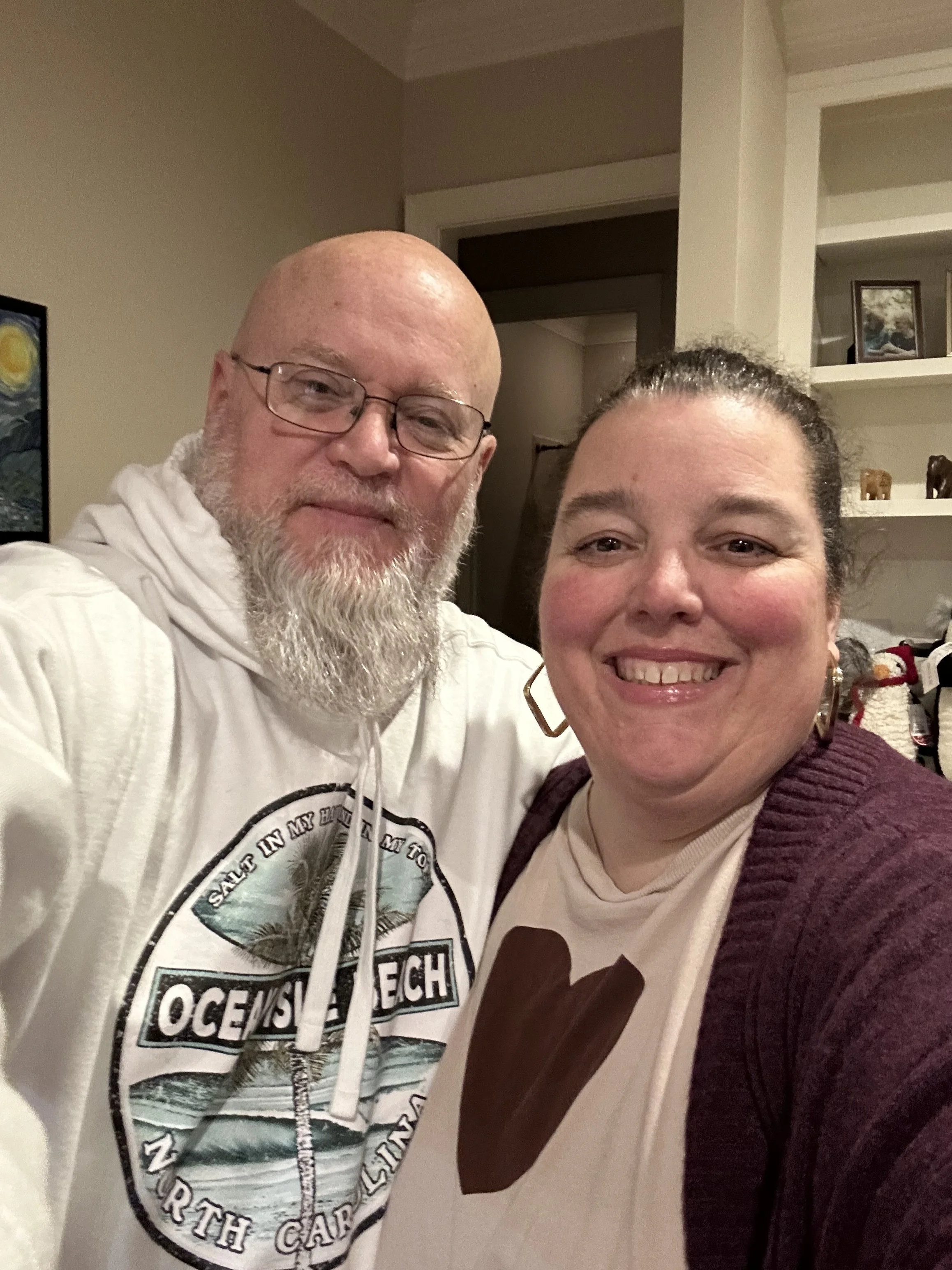 A smiling man and woman taking a selfie together inside a home, with shelves and artwork visible in the background.