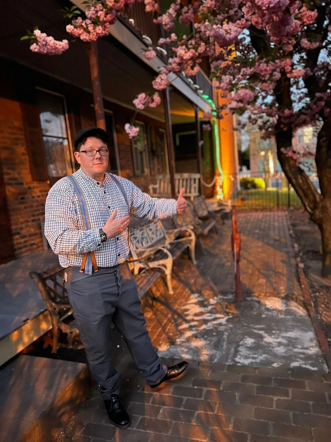 A man wearing glasses, a black cap, a checkered shirt, suspenders, gray pants, and black shoes, standing outside near a flowering pink tree and a brick building at sunset.