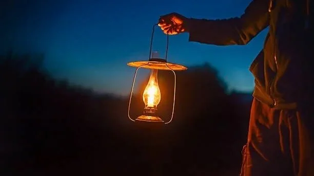 Person holding a lantern during dusk or night, with dark sky and landscape in the background.