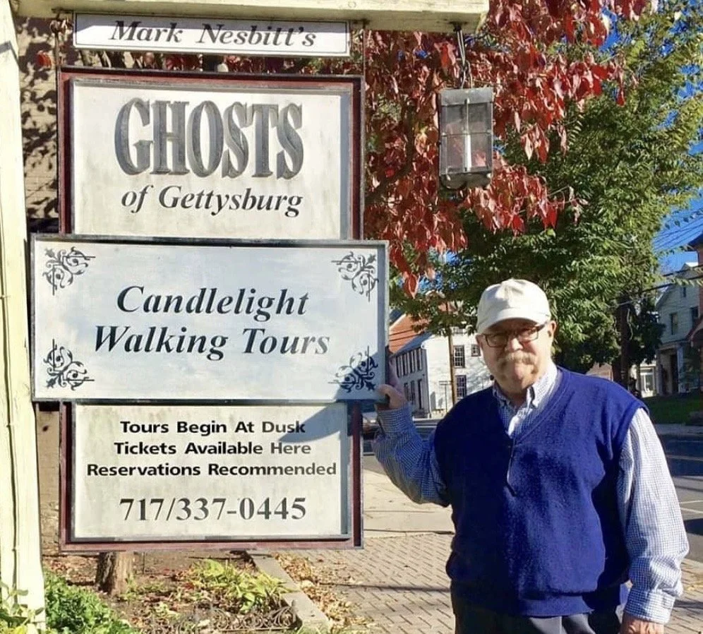 A man with glasses and a white cap standing next to a sign for candlelight walking tours of Gettysburg. The sign is placed on a sidewalk with red and green trees in the background.