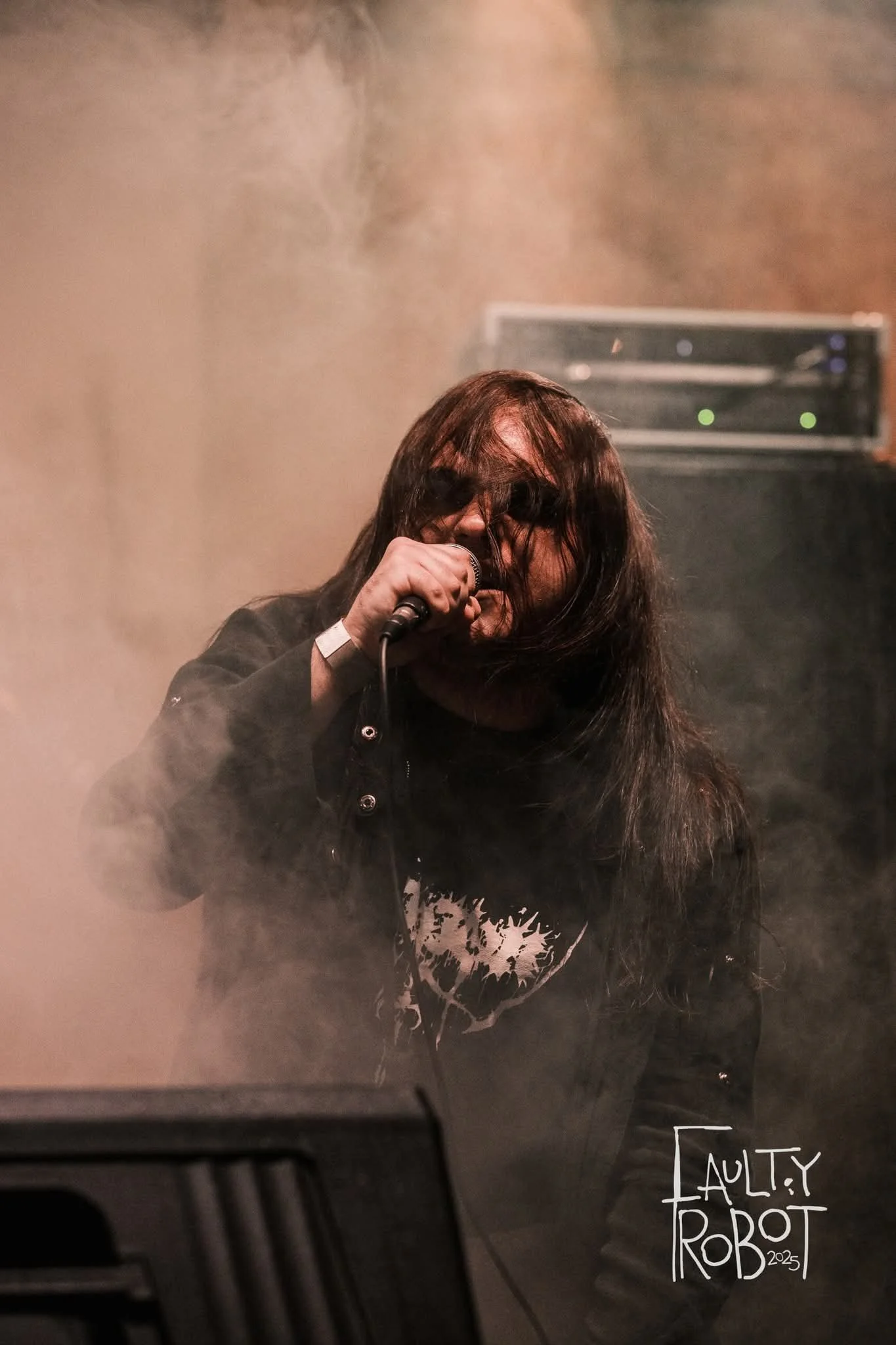 A long-haired person singing into a microphone on stage with smoke and stage equipment in the background.