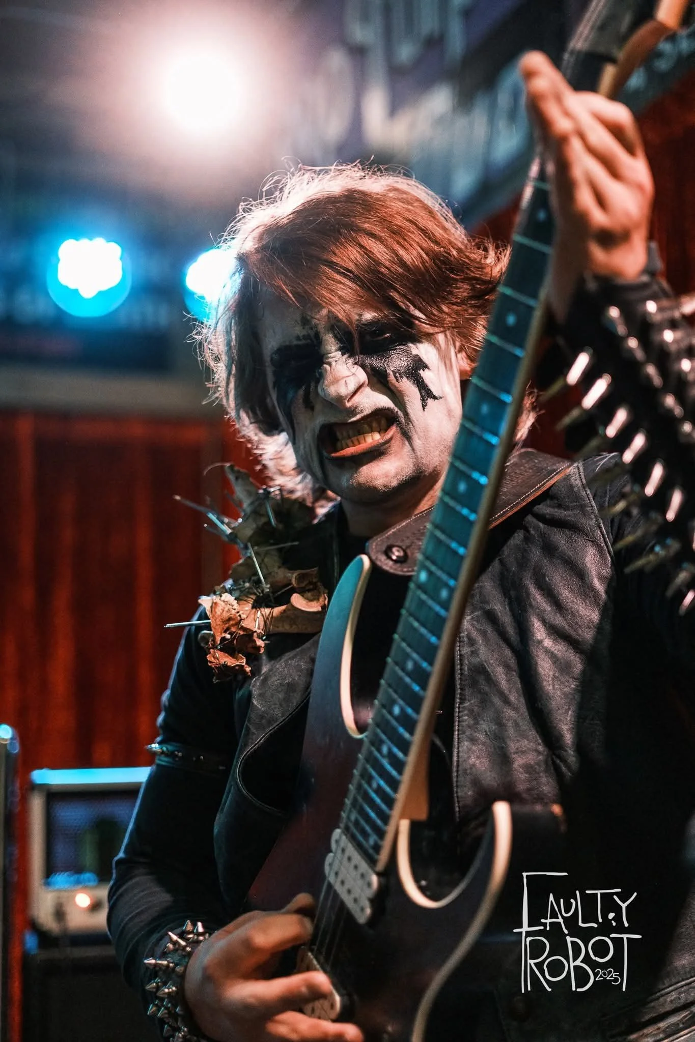 A musician with face paint and devil horns playing an electric guitar on stage with blue and white lights in the background.