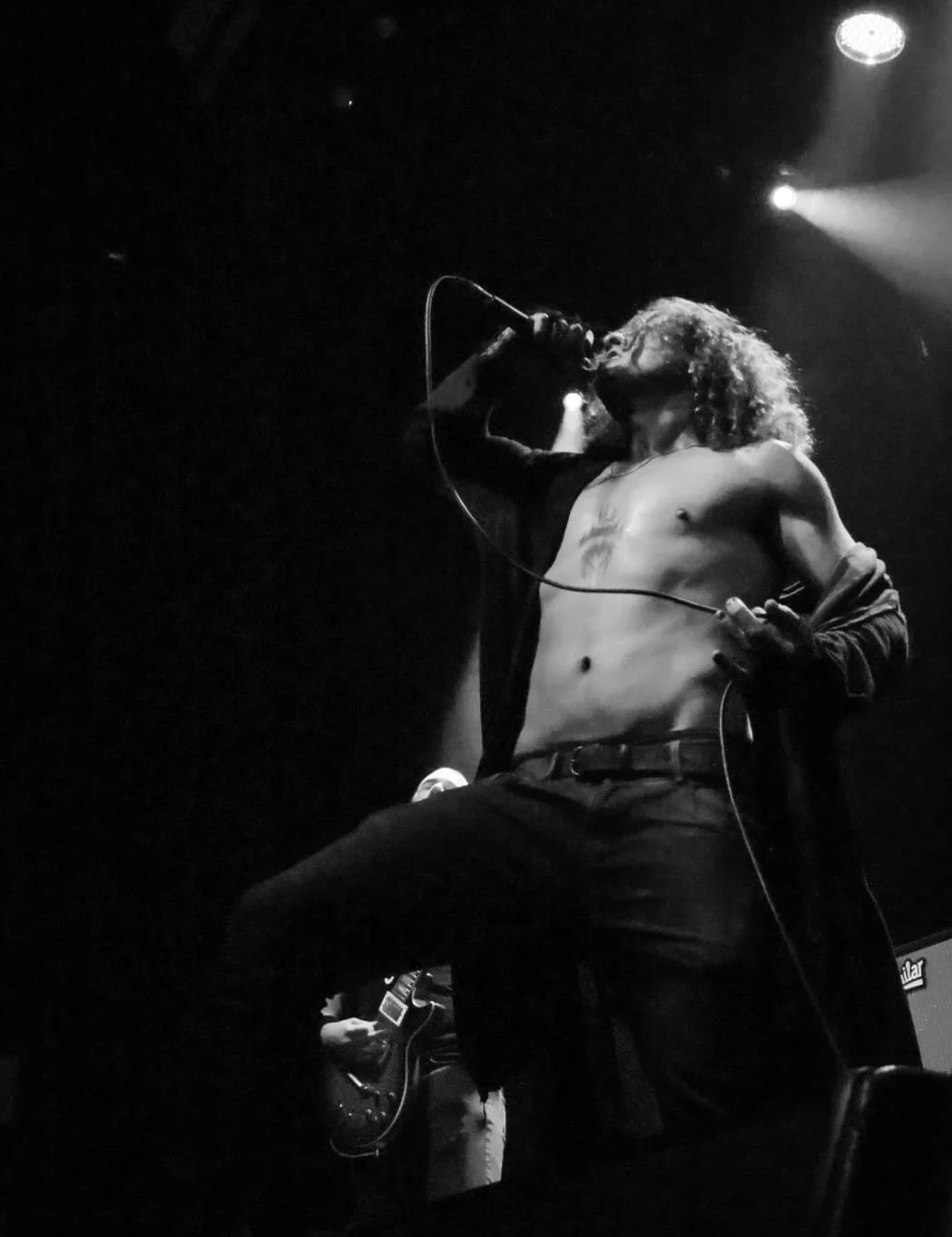 A male singer with curly hair is performing on stage, shirtless, holding a microphone and kneeling, with a guitarist in the background.
