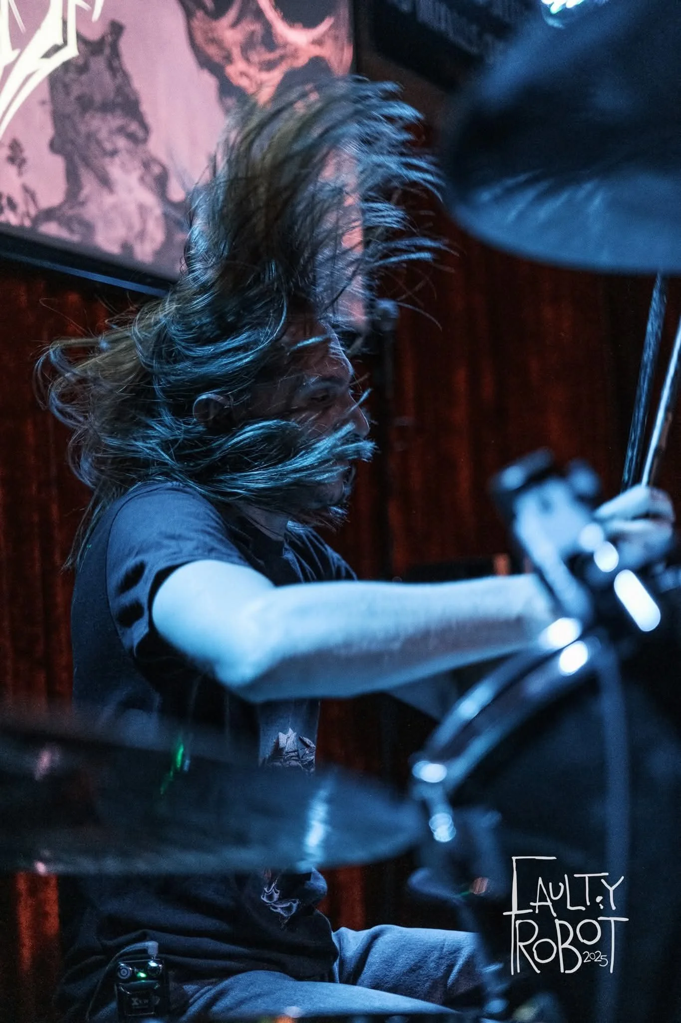 A drummer with long, flowing hair playing drums during a live performance, with a dark background and stage lighting.