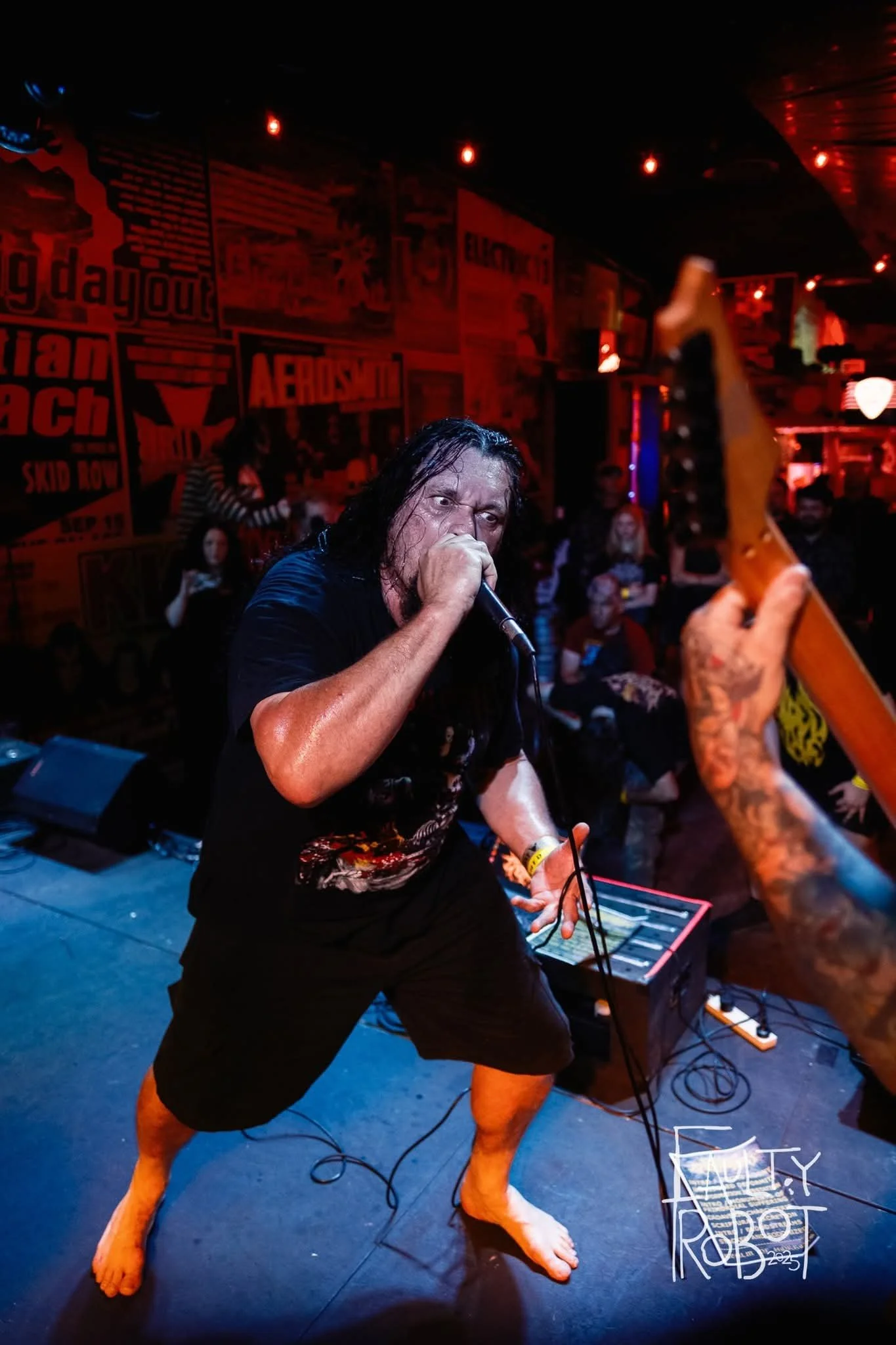 A man with long dark hair and tattoos, barefoot, passionately singing into a microphone on stage in a dimly lit bar or club with red lighting and posters on the wall, holding a guitar.