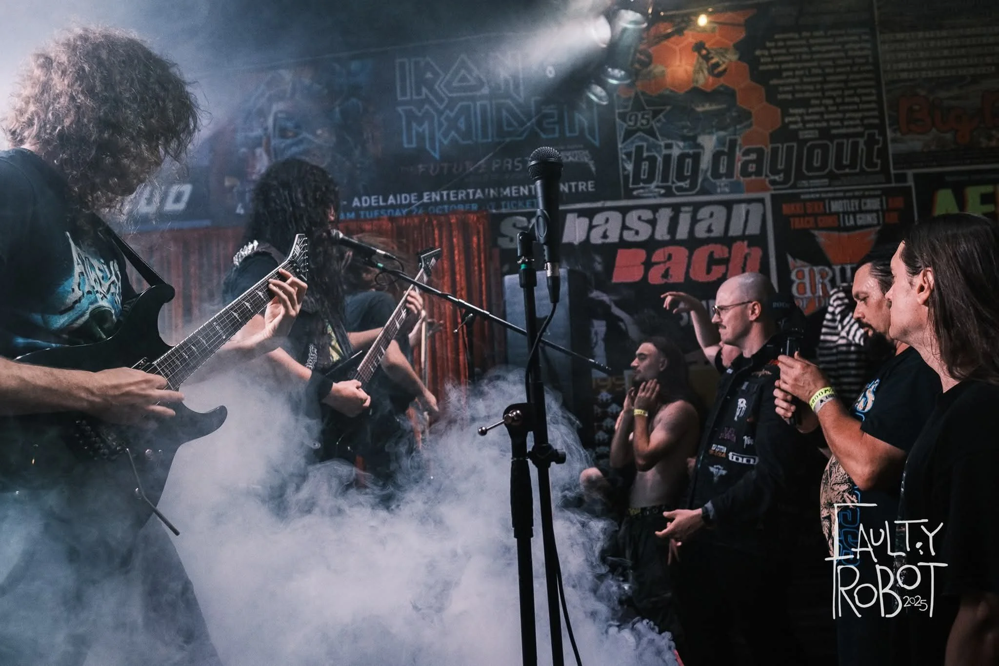 A metal band performing live on stage with fog effects, surrounded by fans and audience members. The stage has colorful posters and banners in the background. Some audience members are looking at the performers, and one is taking a picture.