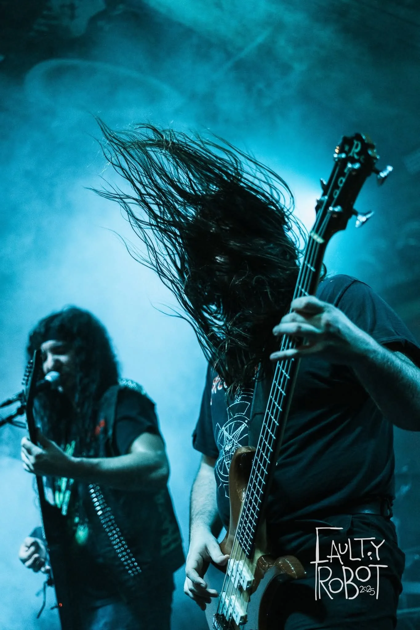 Two musicians performing on stage, one singing into a microphone and the other playing a bass guitar, with their hair flying and blue lighting creating a dynamic concert atmosphere.