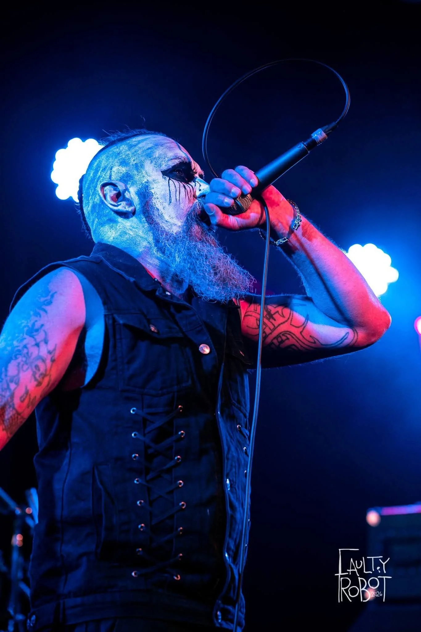 A male vocalist with a painted face and Tattoos singing into a microphone on stage with blue and purple lighting.