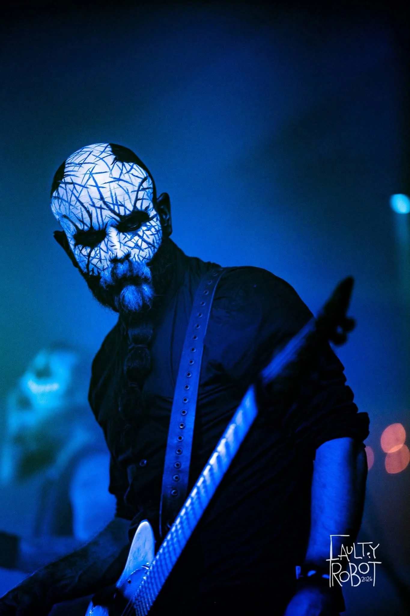A person with a painted face and elaborate makeup, dressed in black, holding a guitar, performing in a dark, blue-lit setting, possibly at a concert or Halloween event.