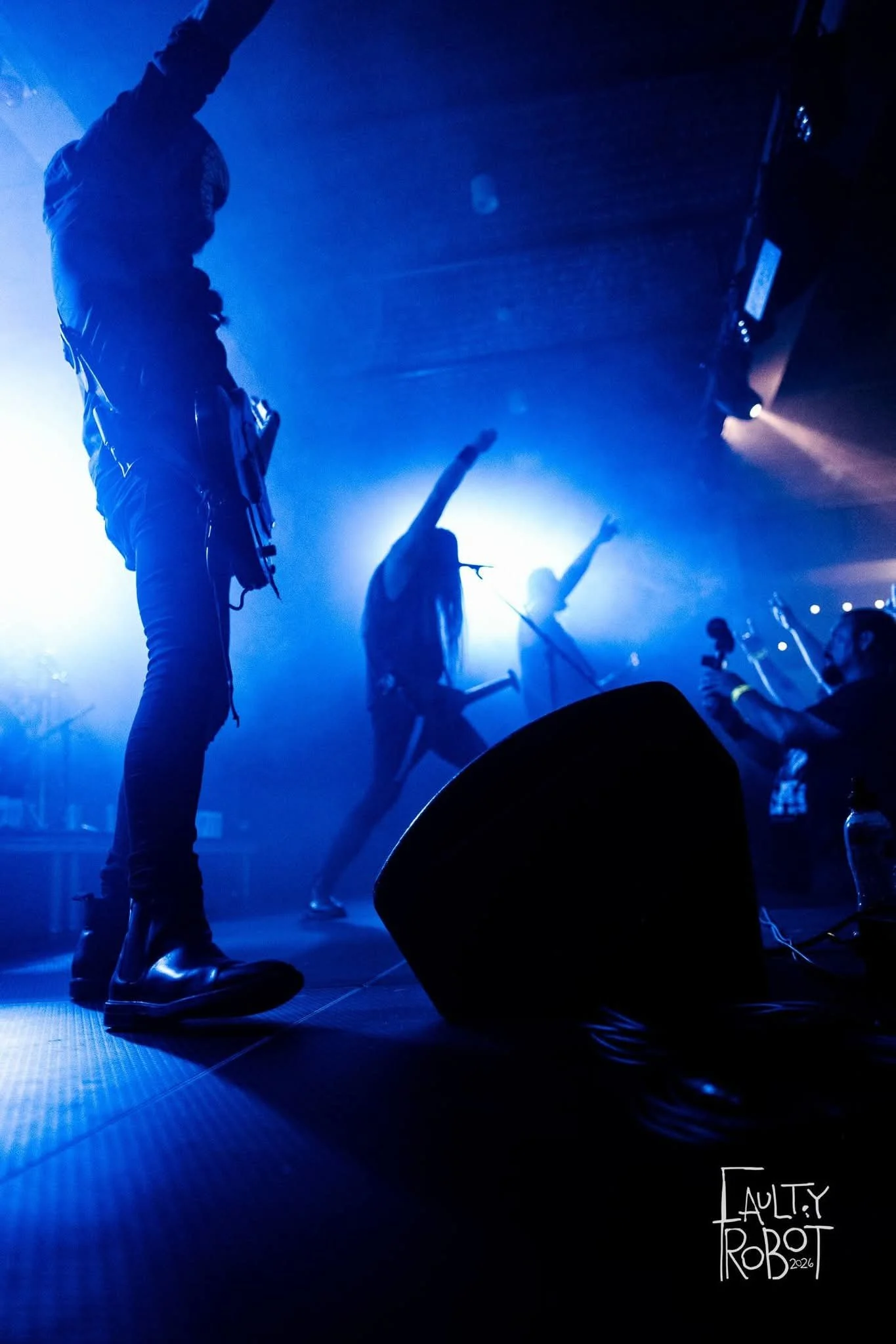Silhouettes of musicians performing on stage with blue lighting, one with an arm raised, and a person with a camera in the audience.