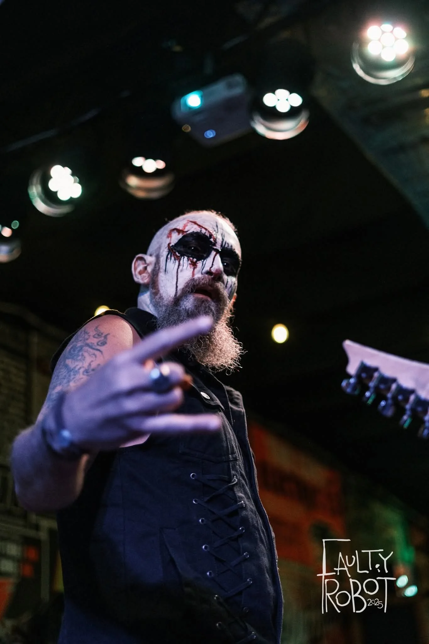 Man with painted face and tattoos gesturing at the camera in a dark indoor venue, with stage lights and equipment overhead, signature 'Faulty Robot 2023' in the corner.