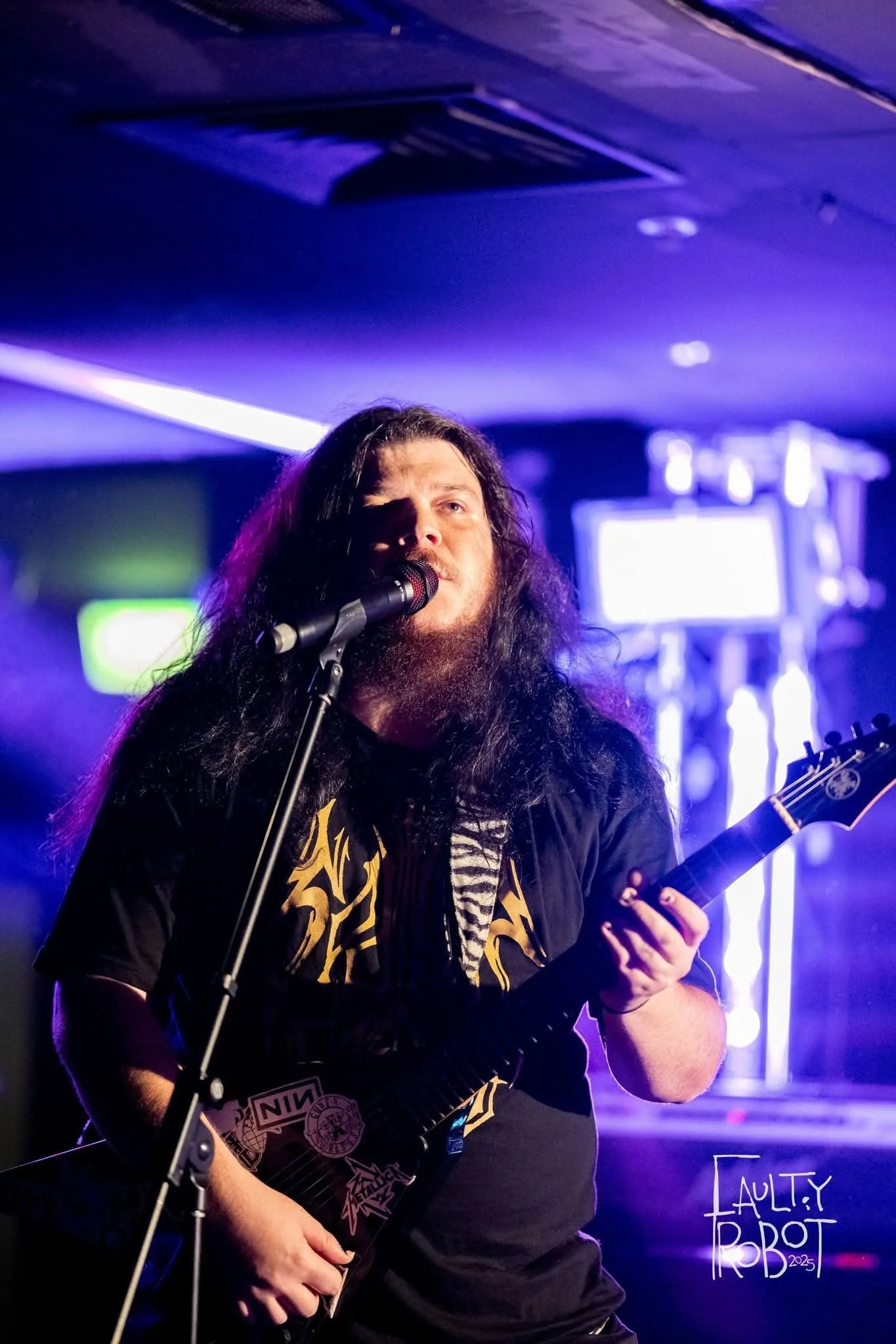A male musician with long dark hair, a beard, and light-colored skin, performing on stage with a black electric guitar and singing into a microphone. The stage is lit with purple and blue lights, and there are screens and equipment in the background.