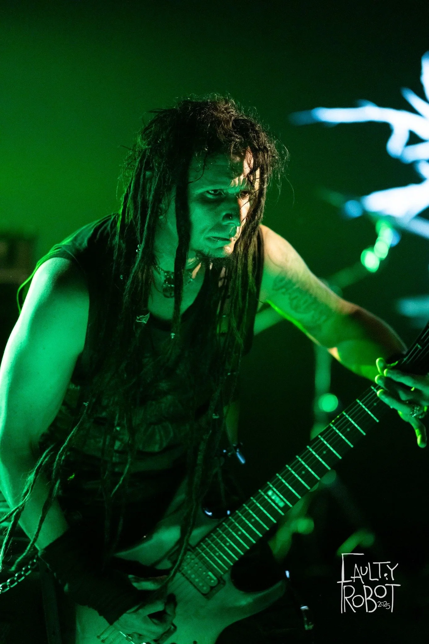 A person with long dreadlocked hair playing an electric guitar on stage, illuminated by green lighting, with a focused and intense expression.