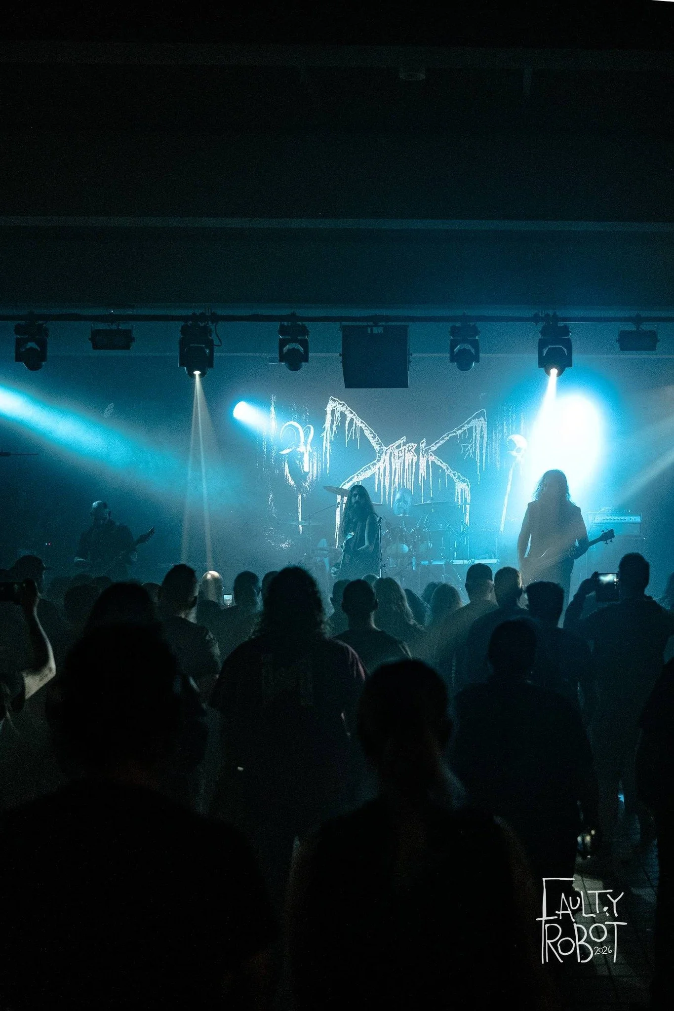 A live band performs on stage with bright blue and white lights illuminating the scene. The audience watches the performance, and a large screen behind the band displays a jagged, abstract graphic. The logo of the photographer, 'Laury Rob 2026,' is i