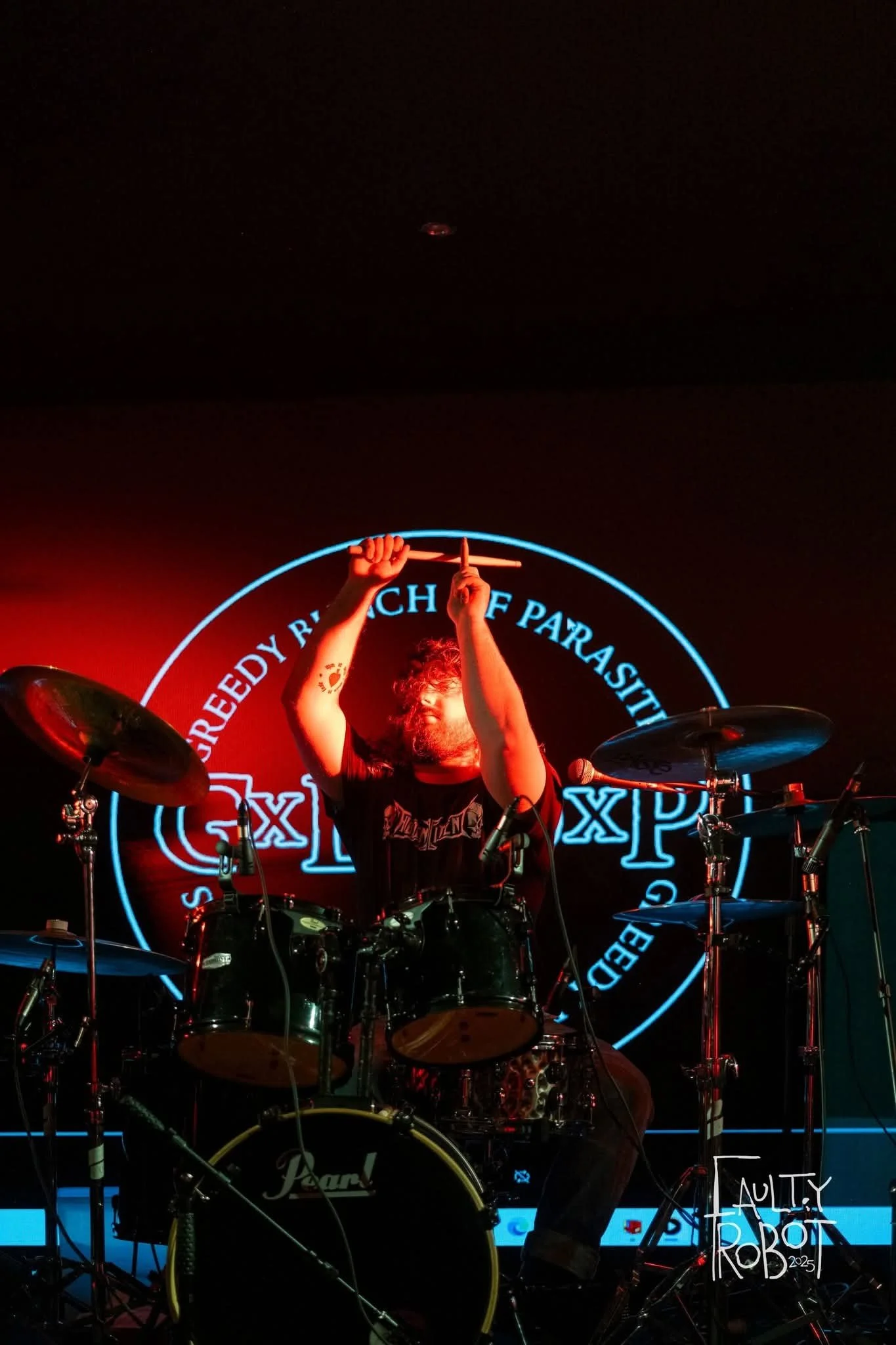 A drummer playing on stage with a neon sign behind him that reads 'Screedy Reach of Parasites' and 'MULTY ROBOT 2025' on a small sign in the bottom right corner.