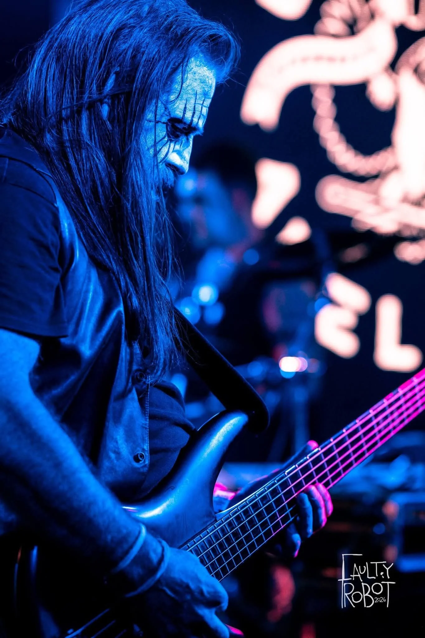 Musician with long hair painted white and clad in dark clothing playing an electric bass guitar on stage, illuminated by blue and pink lights, with a blurred background of a band and stage signage.
