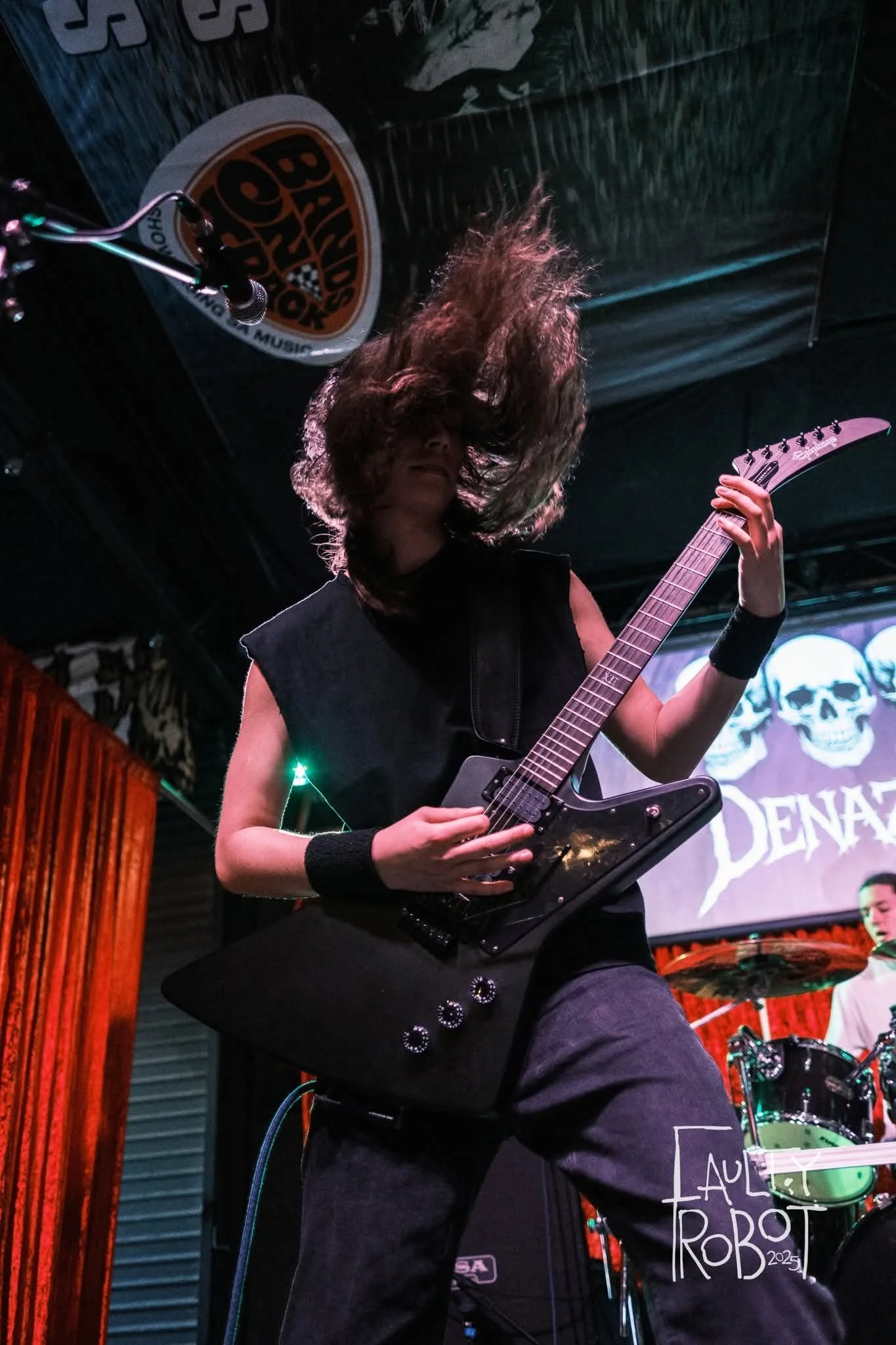 A female guitarist performing on stage with her hair flying, holding an electric guitar in a live music setting with a backdrop displaying skulls and the word 'DENA'.