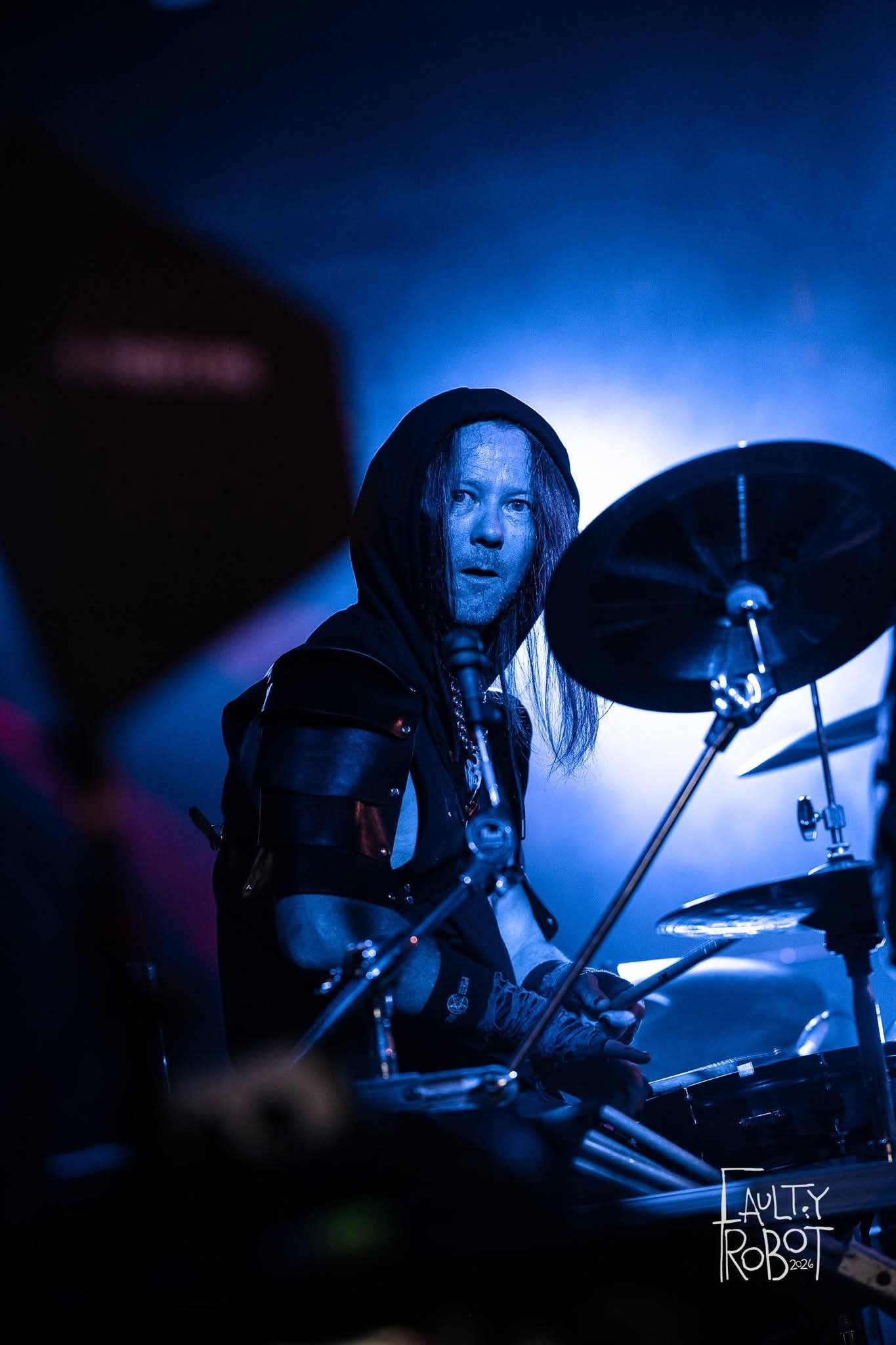 A drummer with long hair, wearing a black hood and clothing, is playing on stage with blue lighting. The photo captures him looking intently at the camera, with a drum set in front and the logo 'Faulty Robot 2026' in the bottom right corner.
